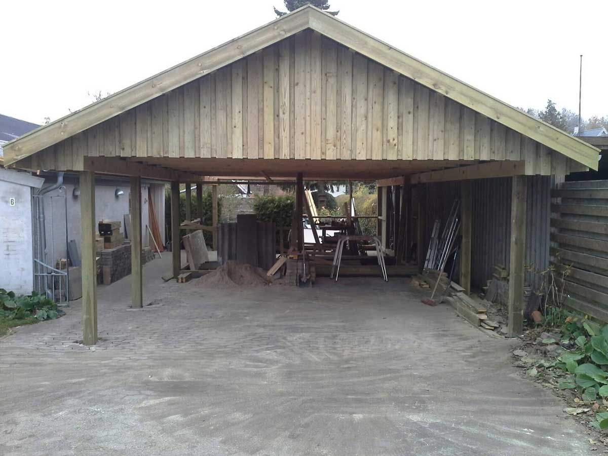 Træcarport med grusindkørsel; værktøj og opbevaring synligt indenfor.