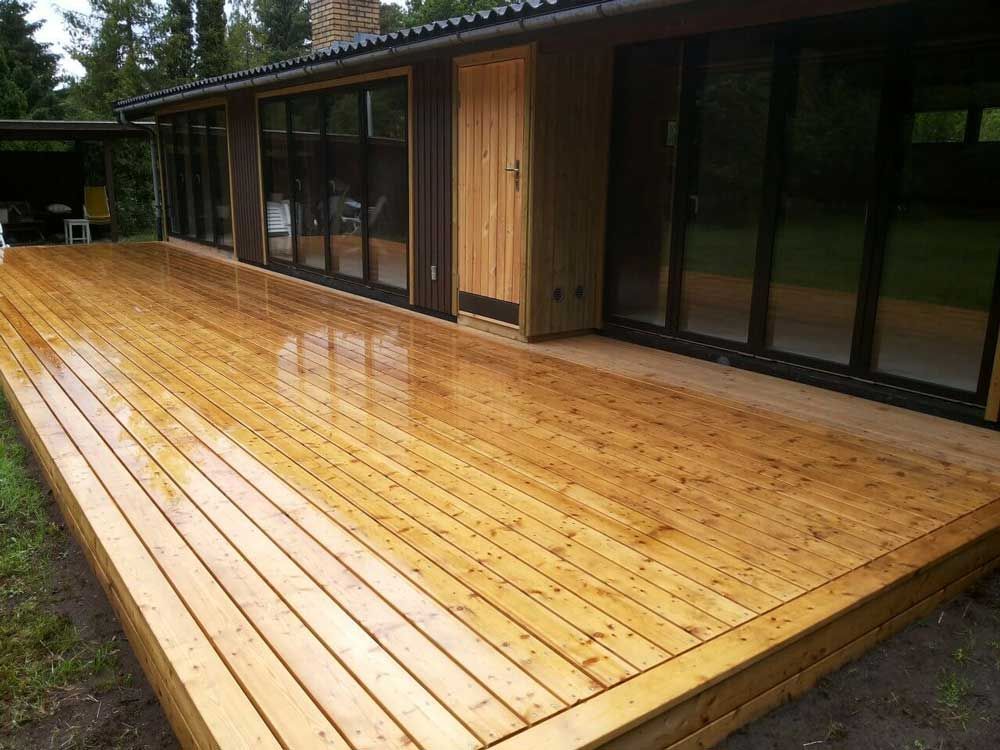 Træterrasse strækker sig fra et brunt hus med store vinduer. Terrassen er våd og udendørs i et græsklædt område.