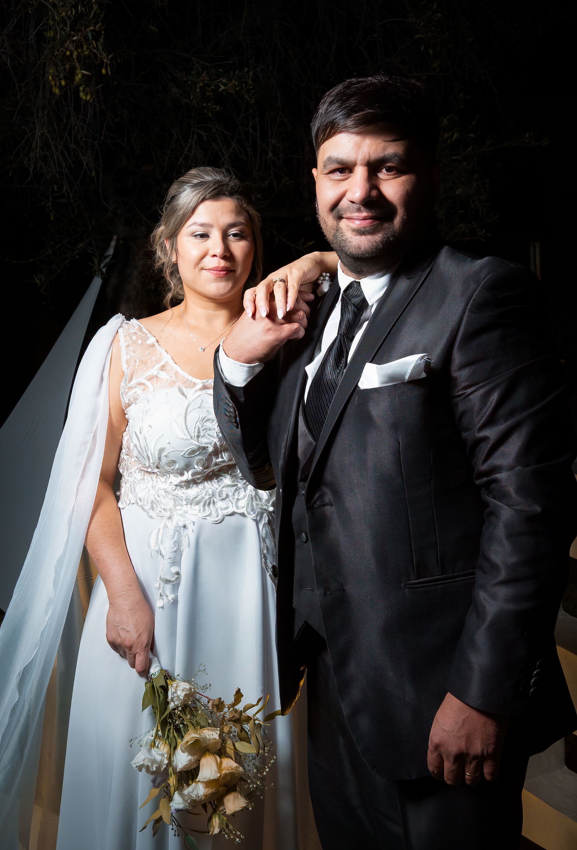 Pareja vestida con atuendo nupcial formal, sonriendo y posando con un fondo oscuro.