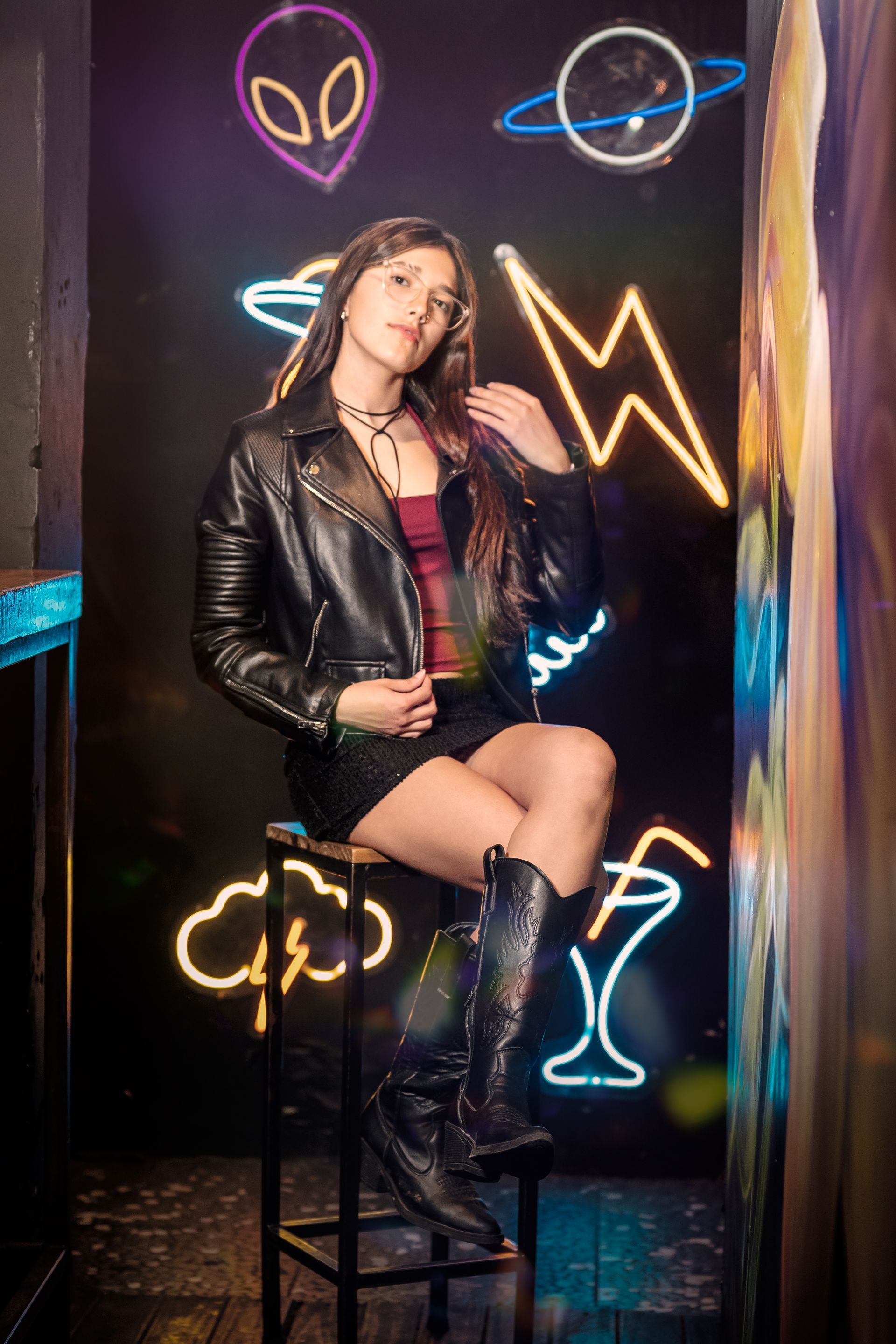 Mujer con chaqueta y botas de cuero negras posando en un taburete entre un mural de neón con temática de alienígenas y rayos.