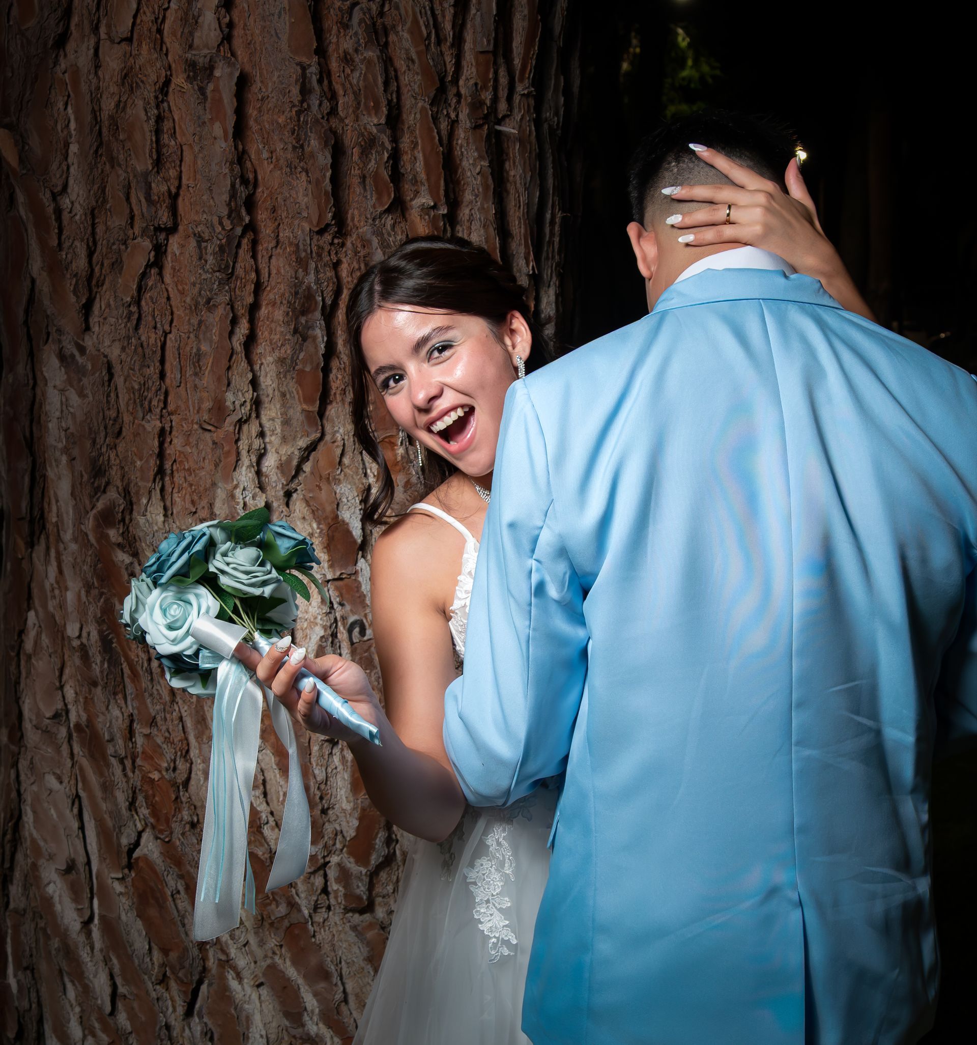 La novia sonríe y sostiene un ramo mientras abraza a su pareja junto al tronco de un árbol por la noche.