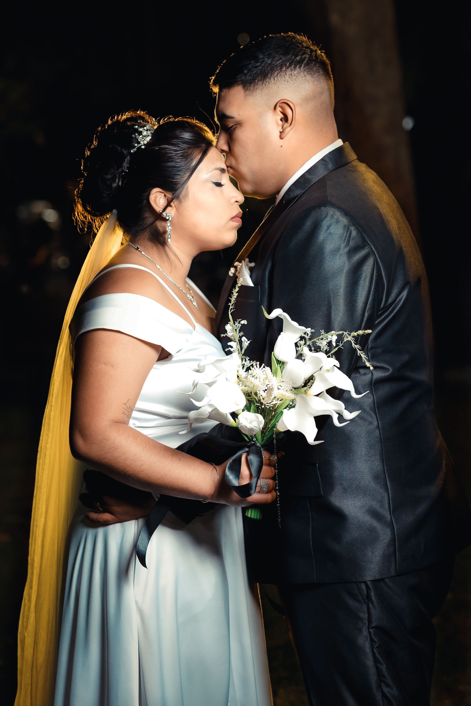 Novios vestidos de boda, de pie muy juntos, sosteniendo un ramo blanco en un ambiente oscuro y romántico.