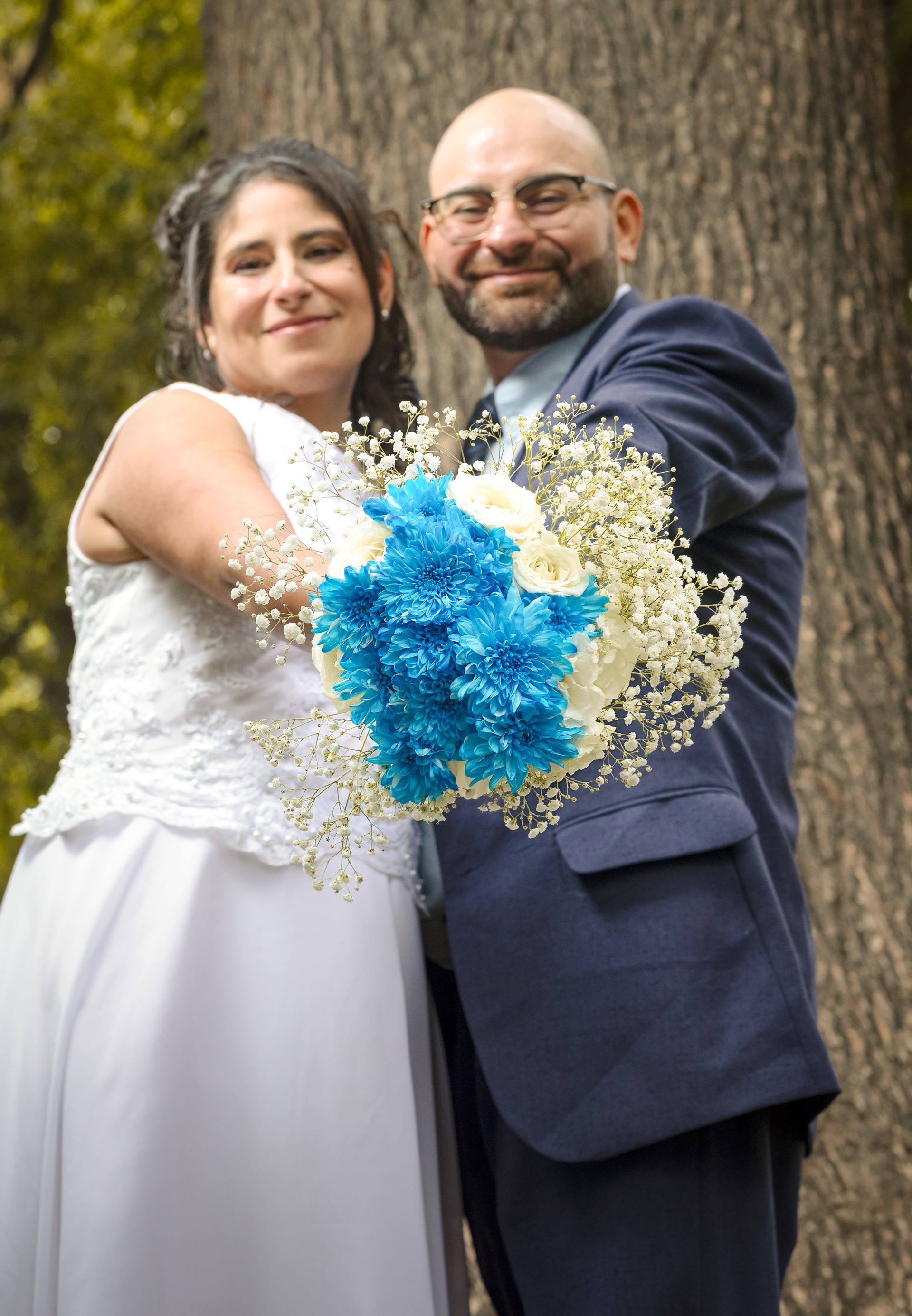 Pareja vestida de novios sosteniendo un ramo azul y blanco al aire libre junto a un árbol.