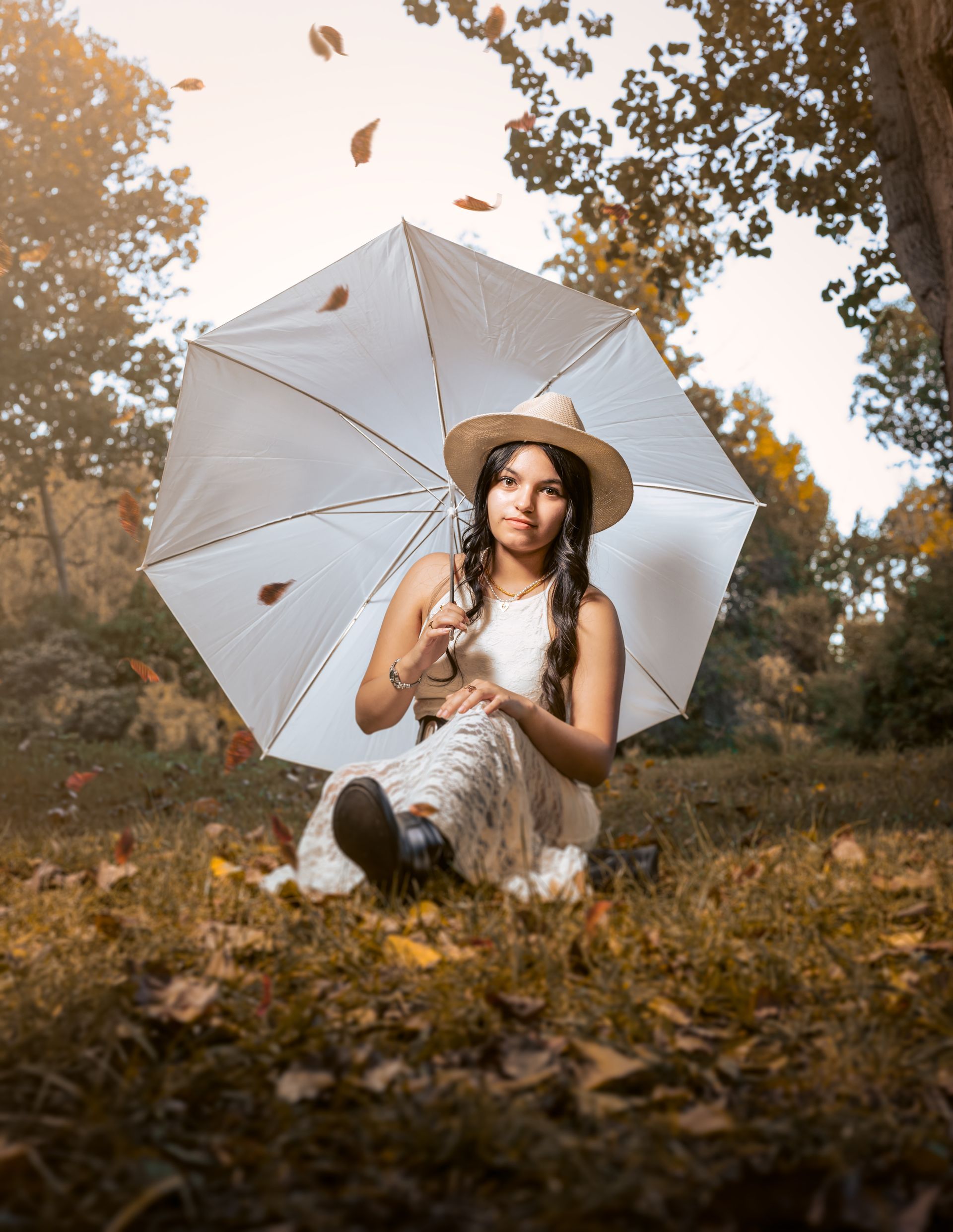 Persona sentada entre hojas otoñales, sosteniendo un paraguas blanco, con sombrero y vestido, en un parque arbolado.