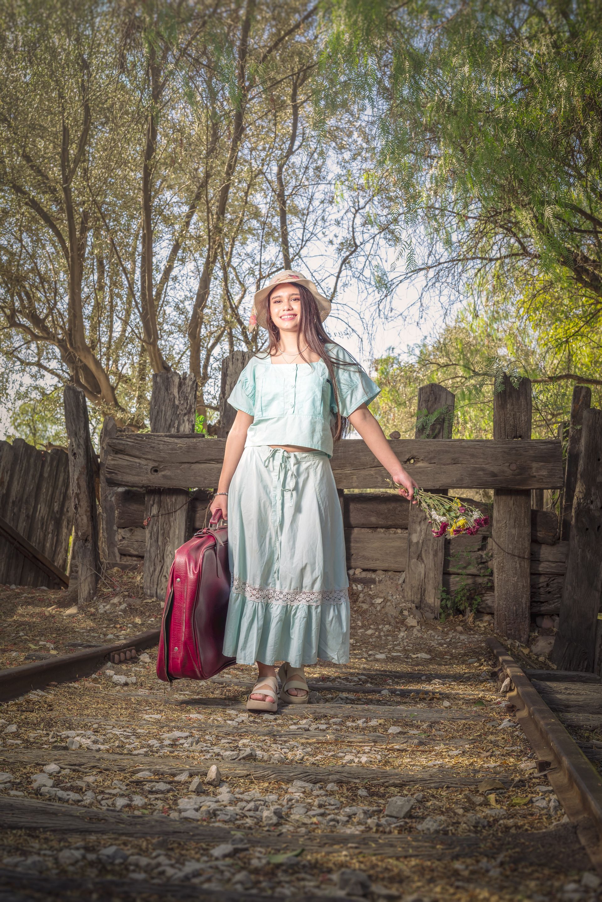 Una persona con un vestido azul claro está de pie sobre un puente rústico junto a un bolso rosa.
