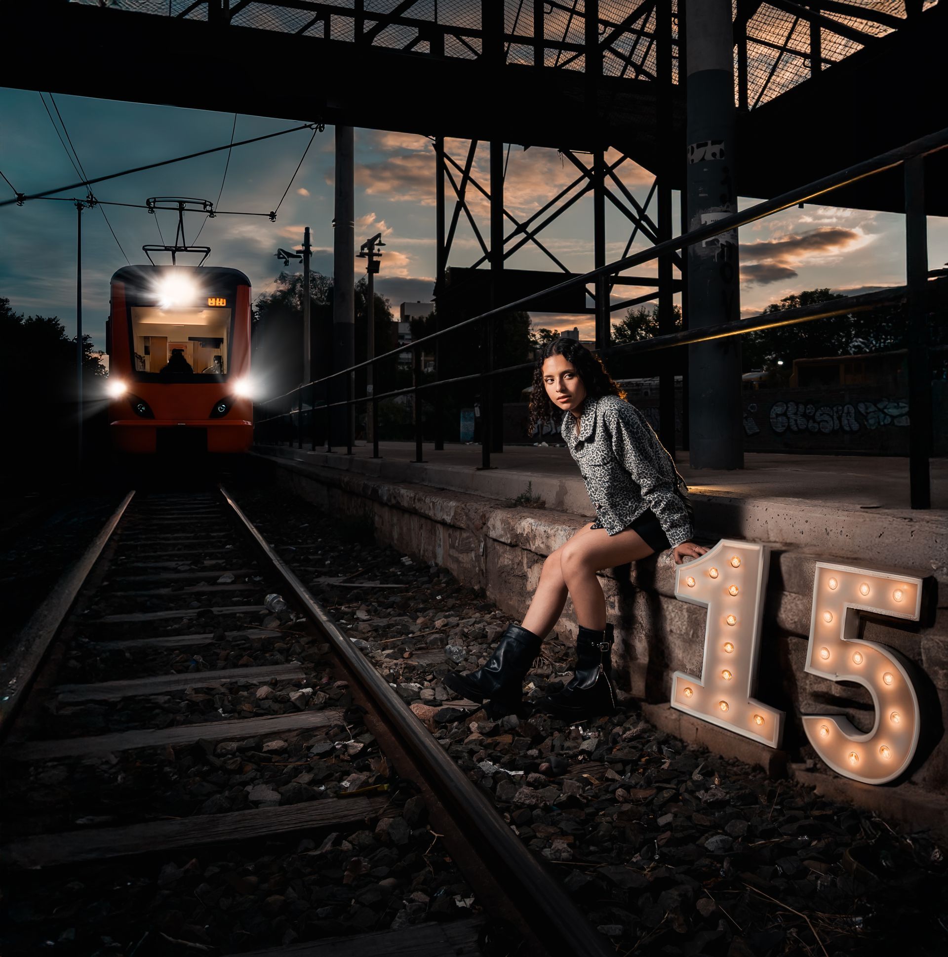 Mujer sentada en las vías del tren al atardecer junto al número 15 iluminado, con un tren que se aproxima al fondo.