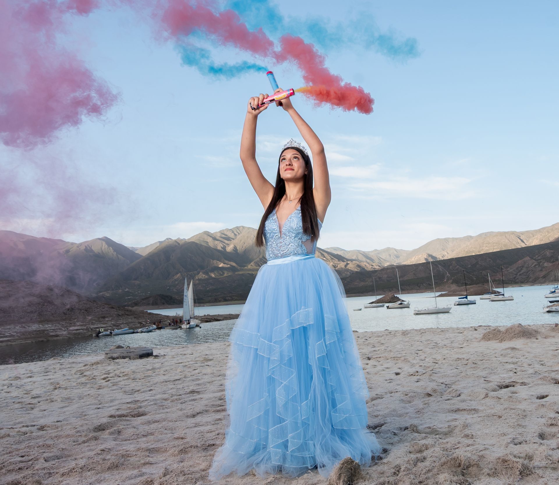 Mujer con vestido azul lanzando bengalas de humo rojo y azul en una playa con montañas y barcos al fondo.