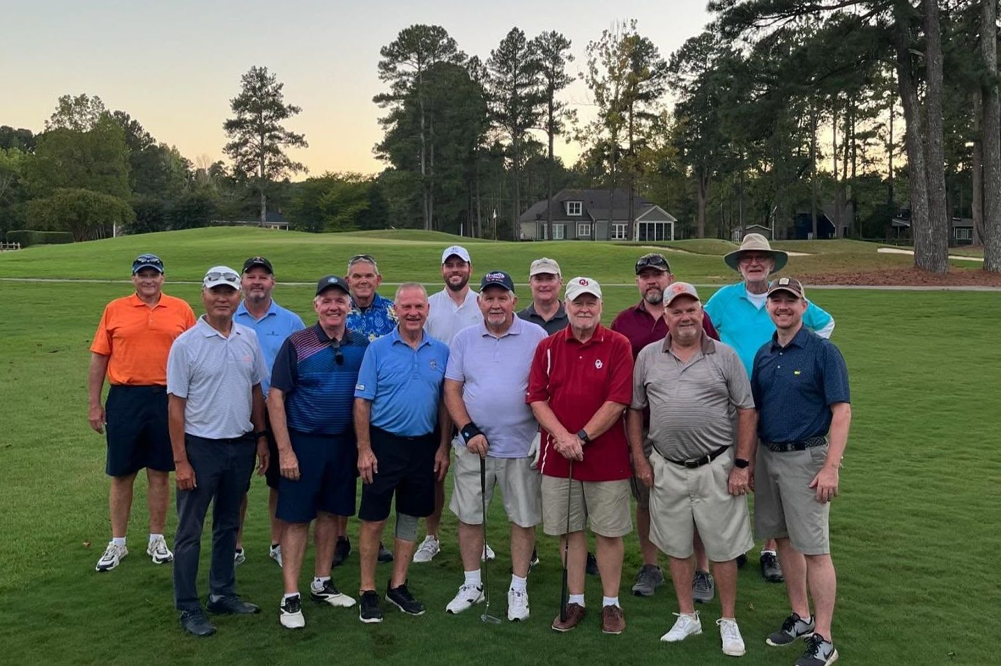 A group of men in golf attire pose on a green, smiling, near trees and a building.