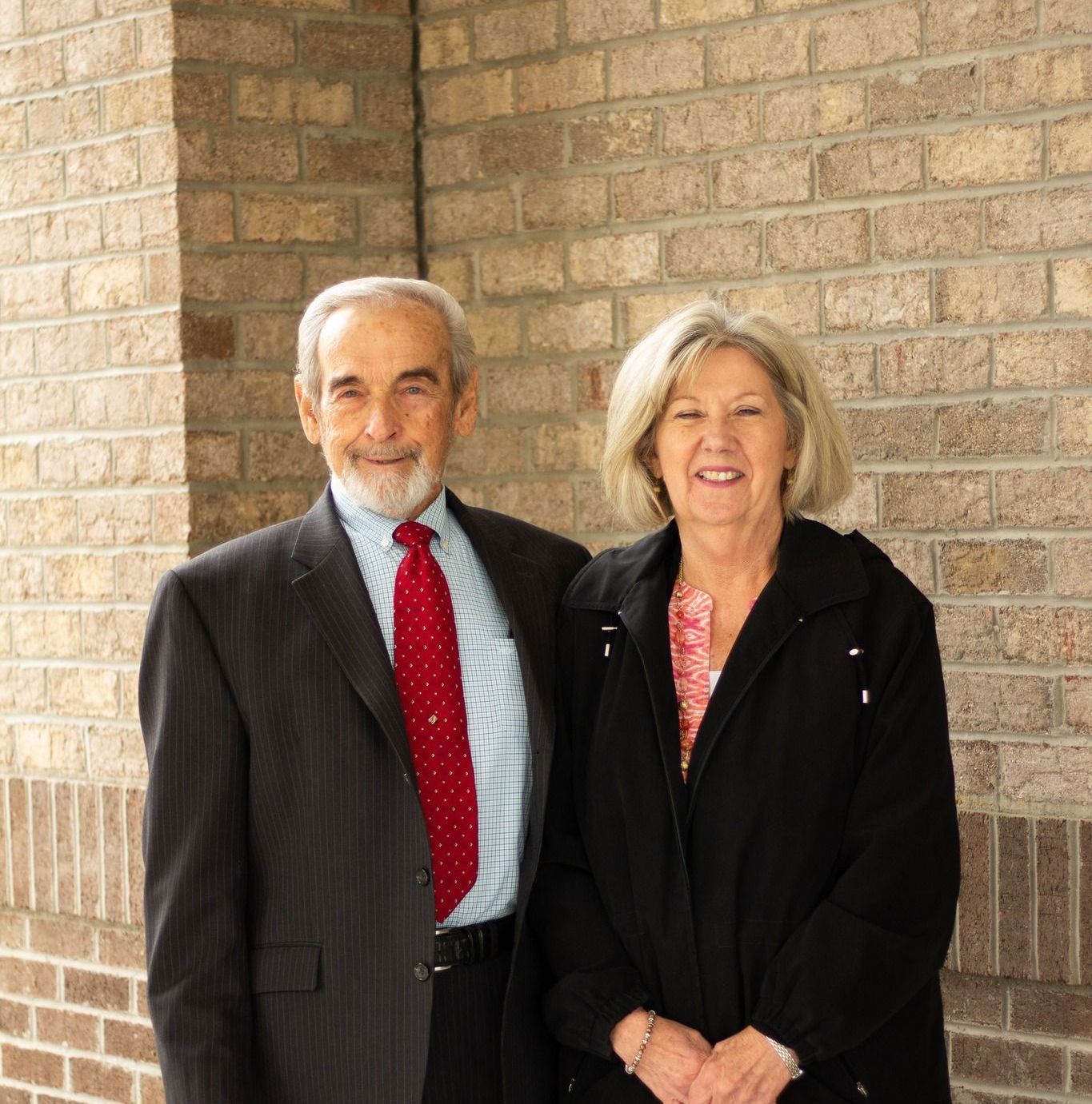Older man in suit and woman in black jacket smiling by brick wall.