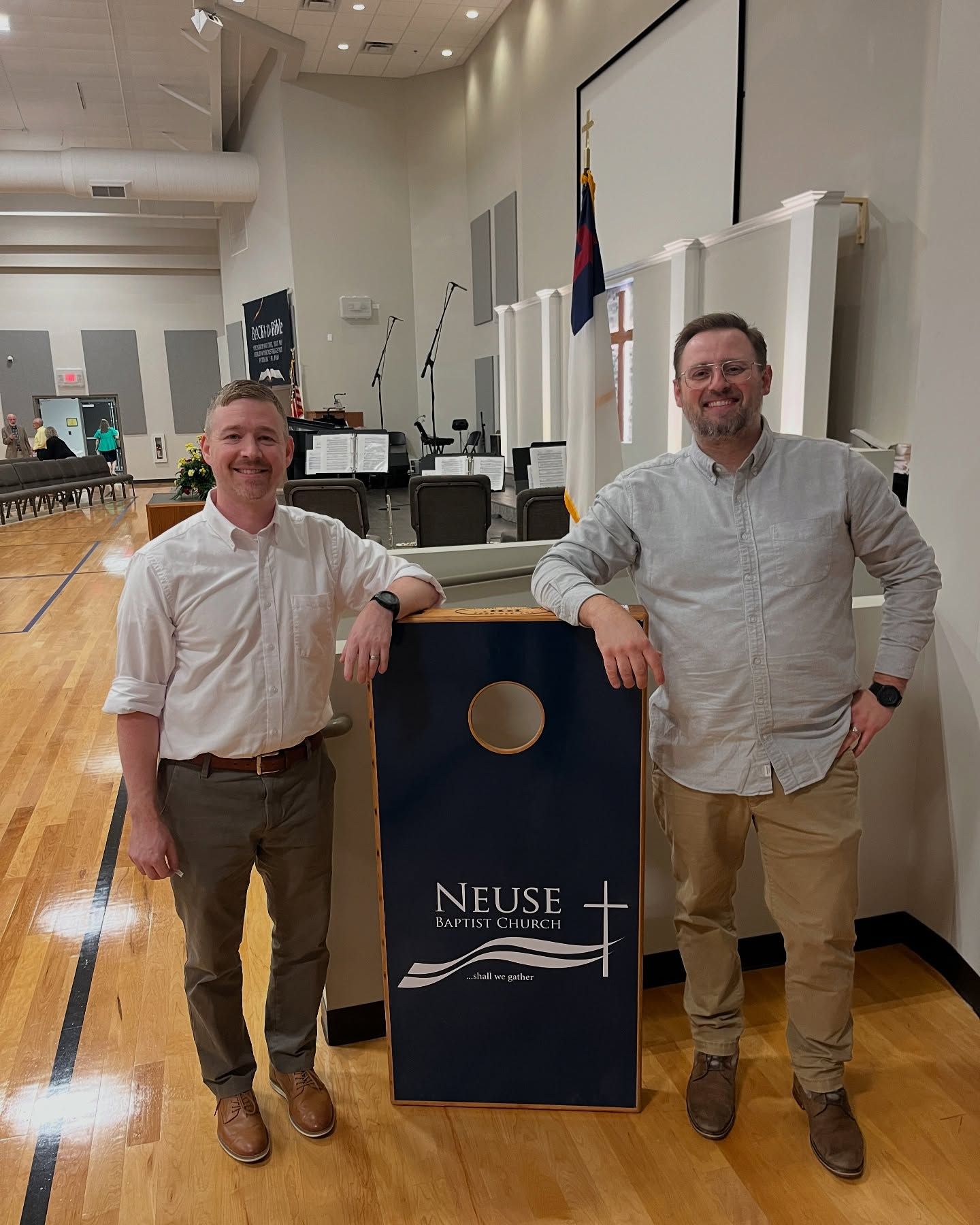 Two men pose with a navy cornhole board. They are in a large room with a wooden floor.