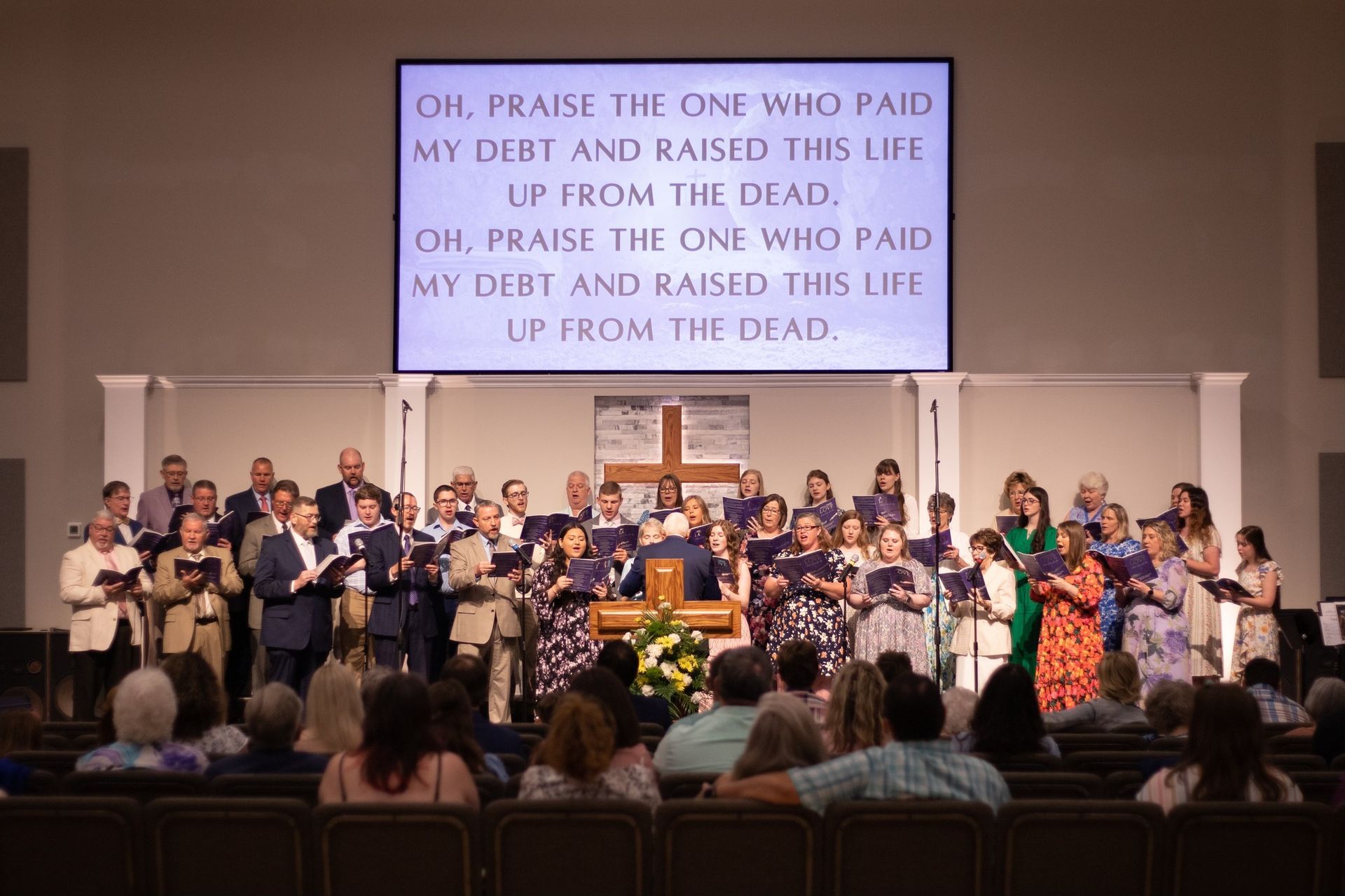 Choir singing in church with lyrics displayed. Audience seated below. Cross visible.