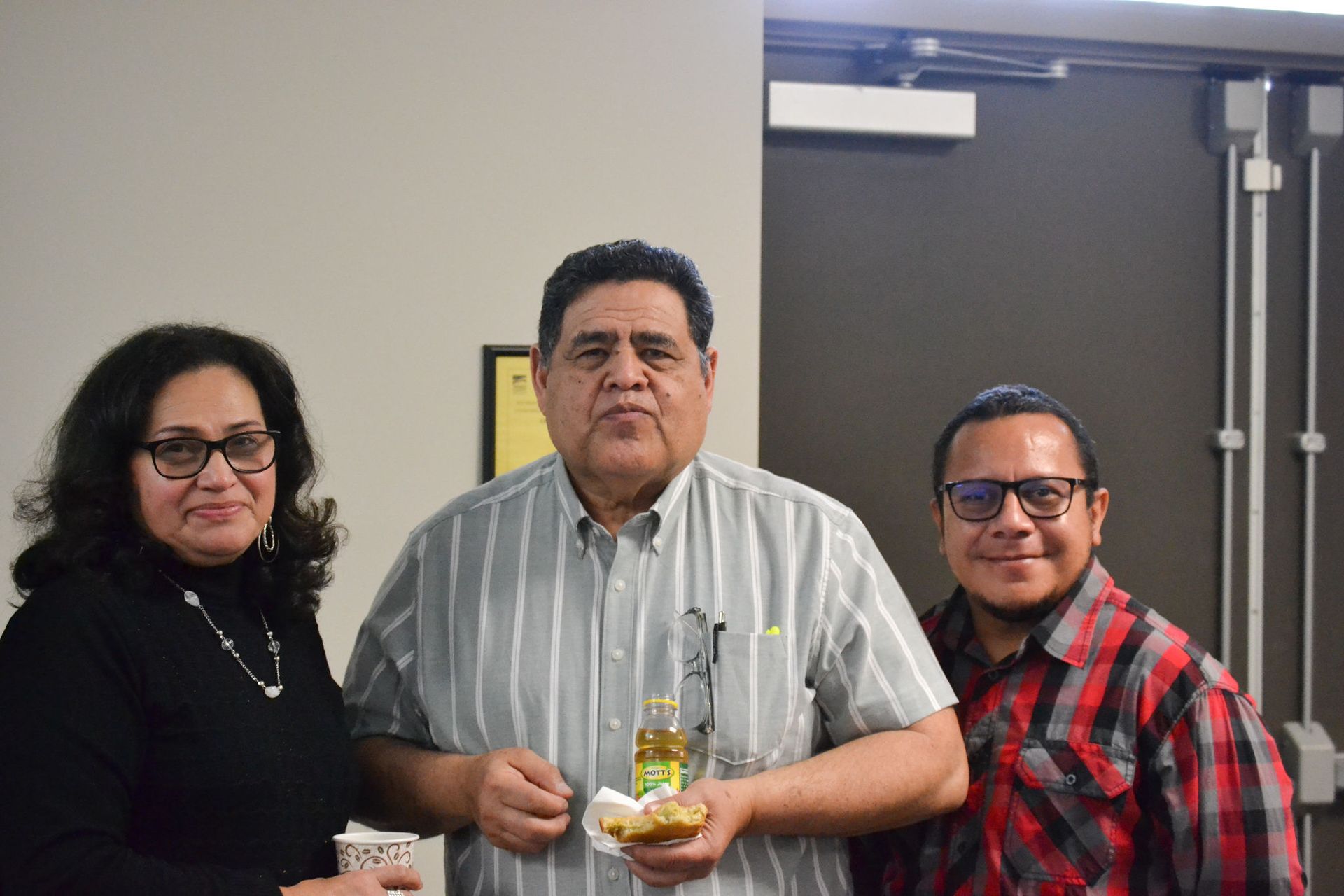 Three people smiling at a gathering; a woman in black, a man with a snack, and a man in a plaid shirt.