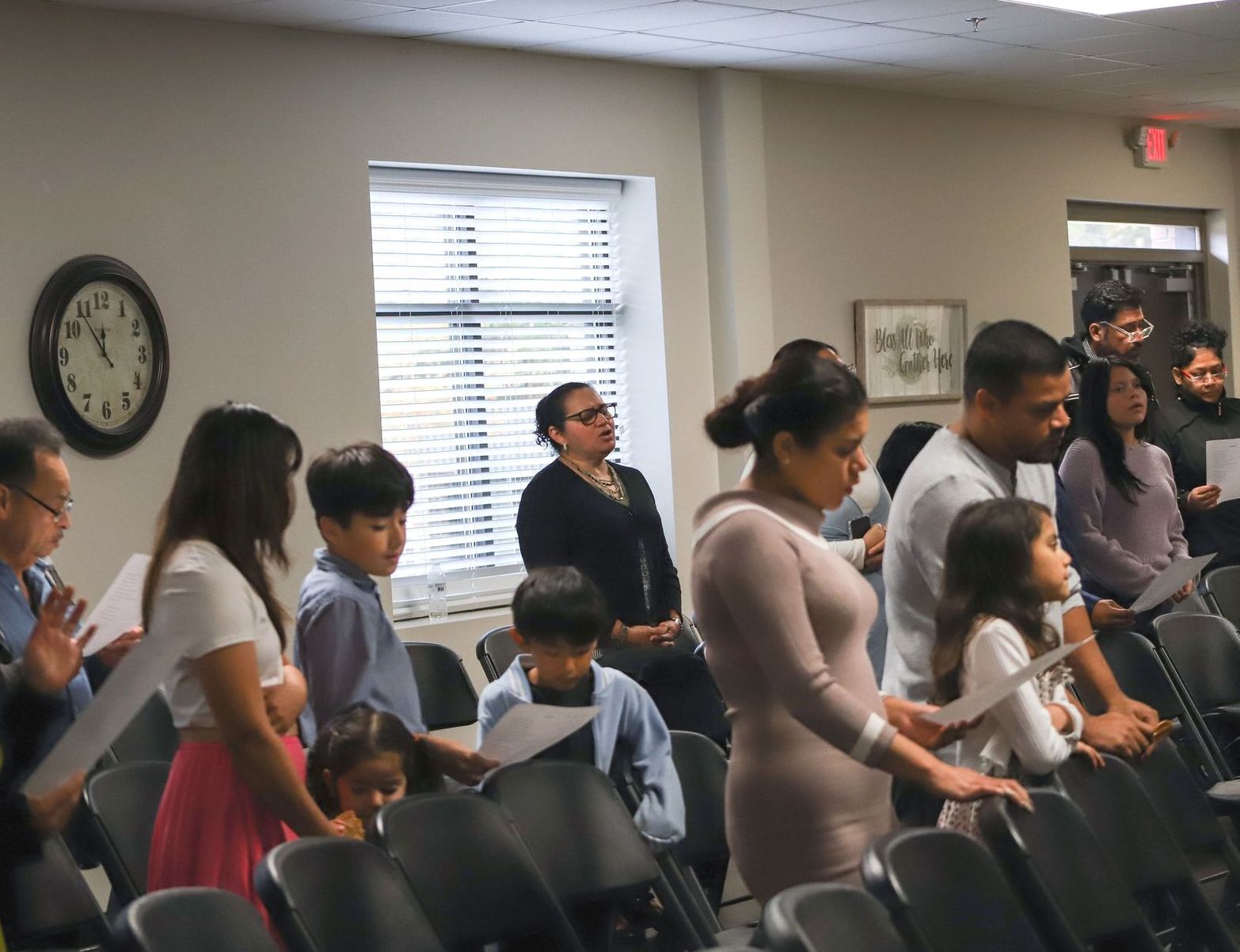 People standing in a room, likely a meeting or church, some with papers, others with hands clasped.