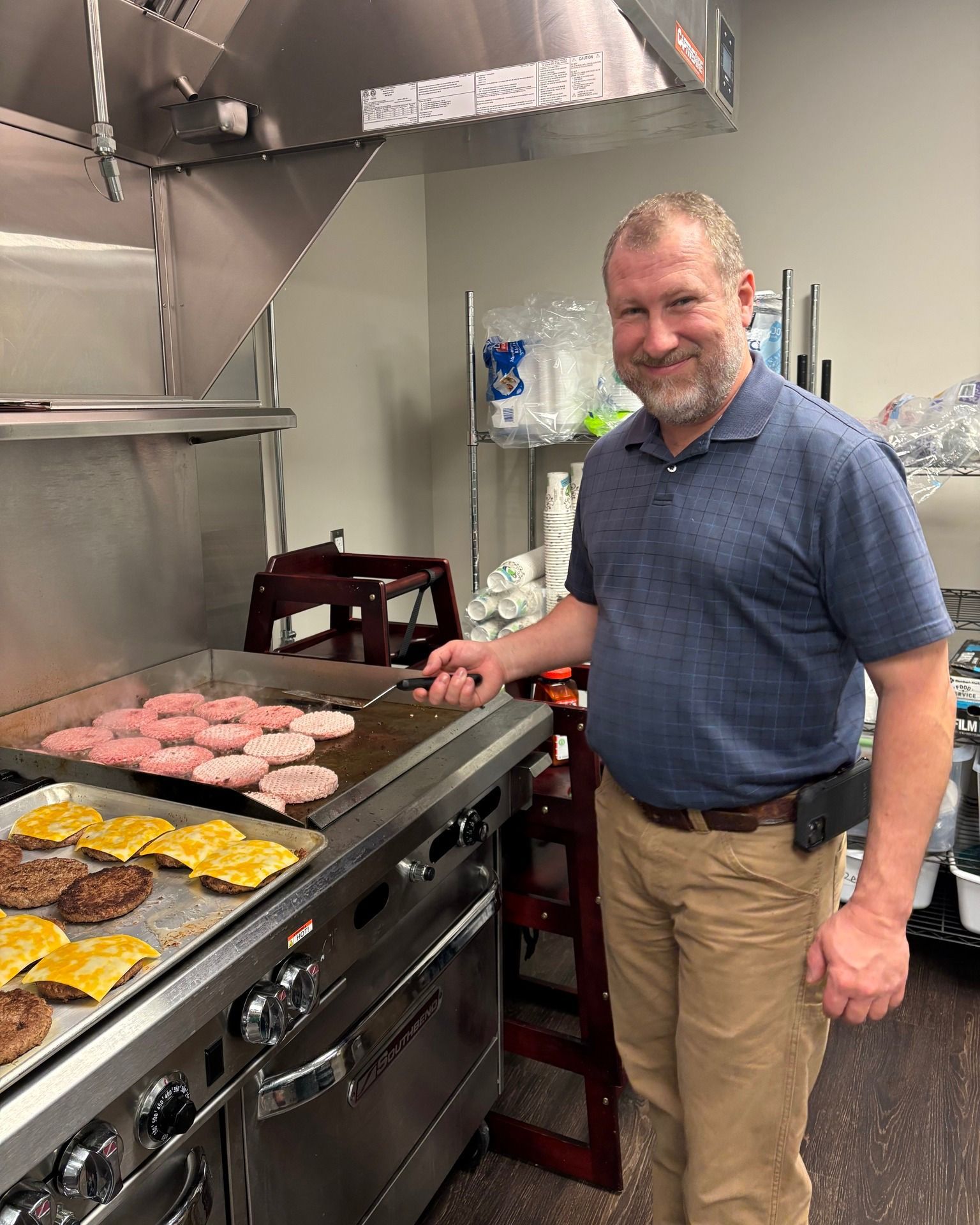 Man grilling burgers on a flat top. He smiles, wearing a blue shirt and tan pants, in a kitchen.