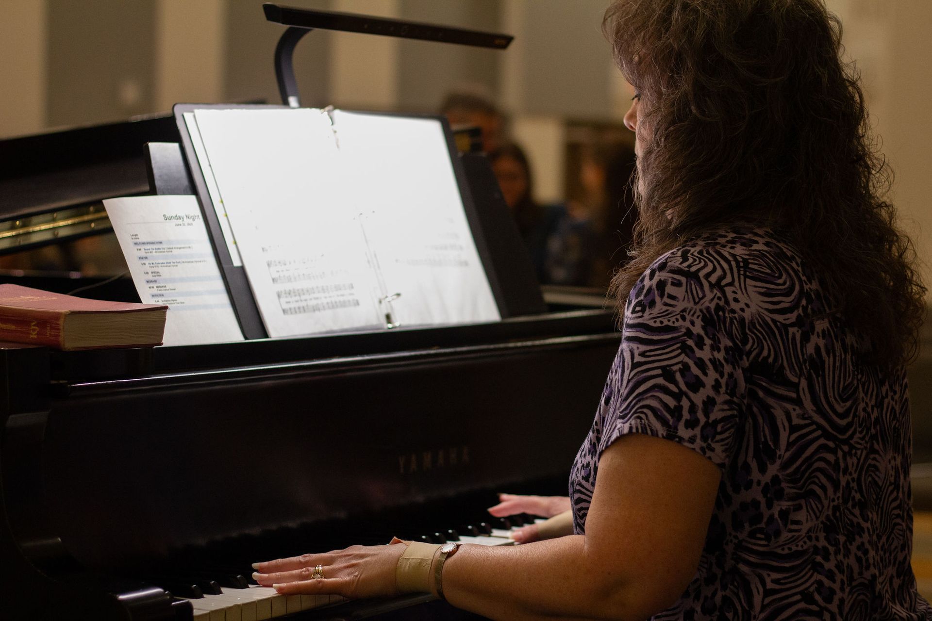Woman playing piano, reading sheet music. Indoor setting with a light focused on the music.