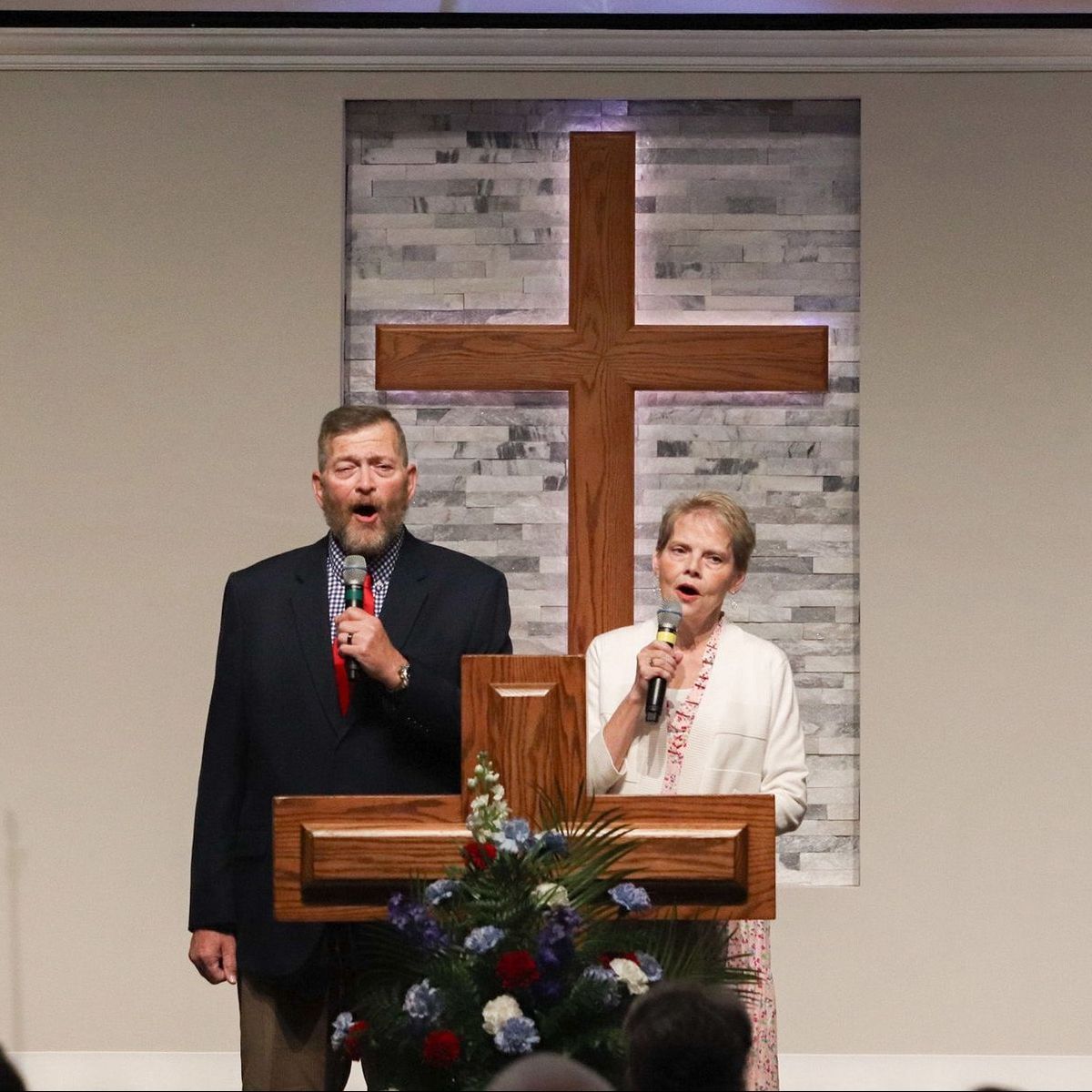 Man and woman singing into microphones at a wooden podium with a cross backdrop.