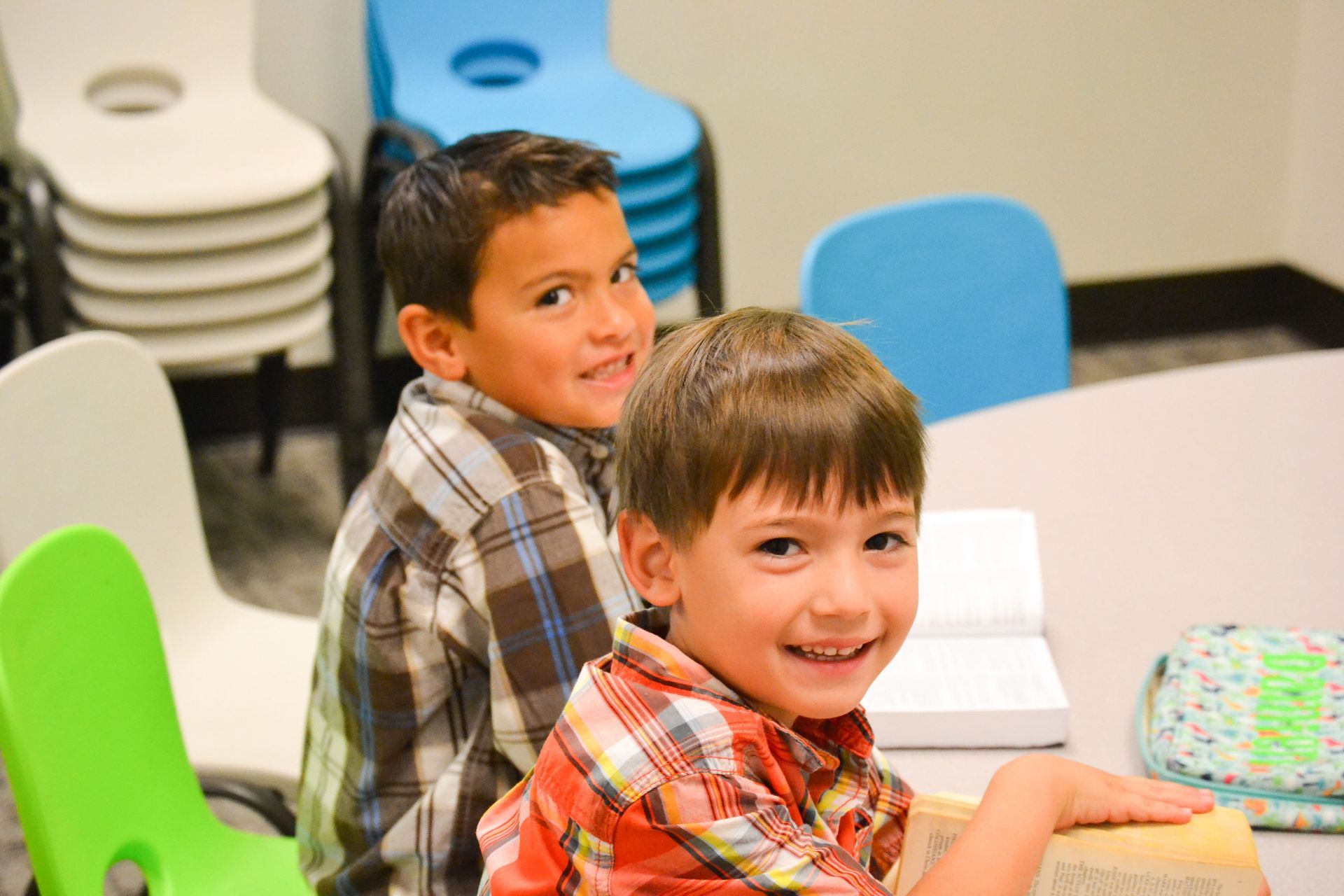 Two smiling boys looking at the camera at a table. One wears an orange plaid shirt. Other, blue plaid.