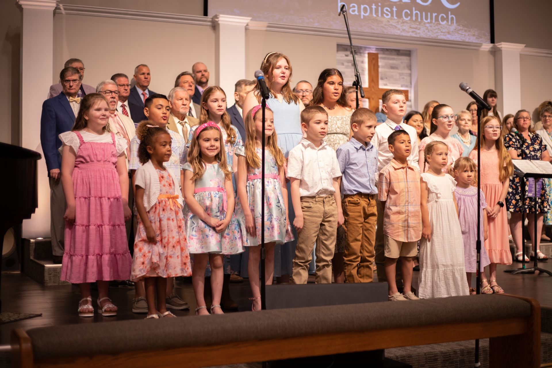 Choir singing in a church; children and adults dressed in light-colored clothing; wooden cross on the right.