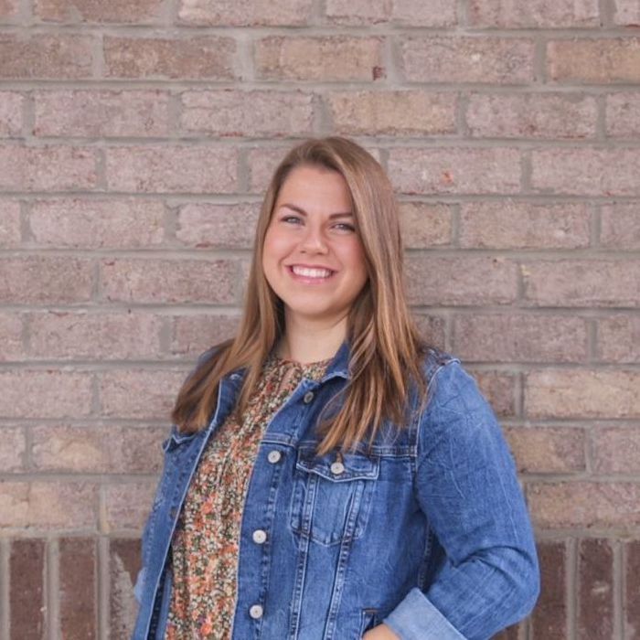 A woman in a denim jacket is standing in front of a brick wall.