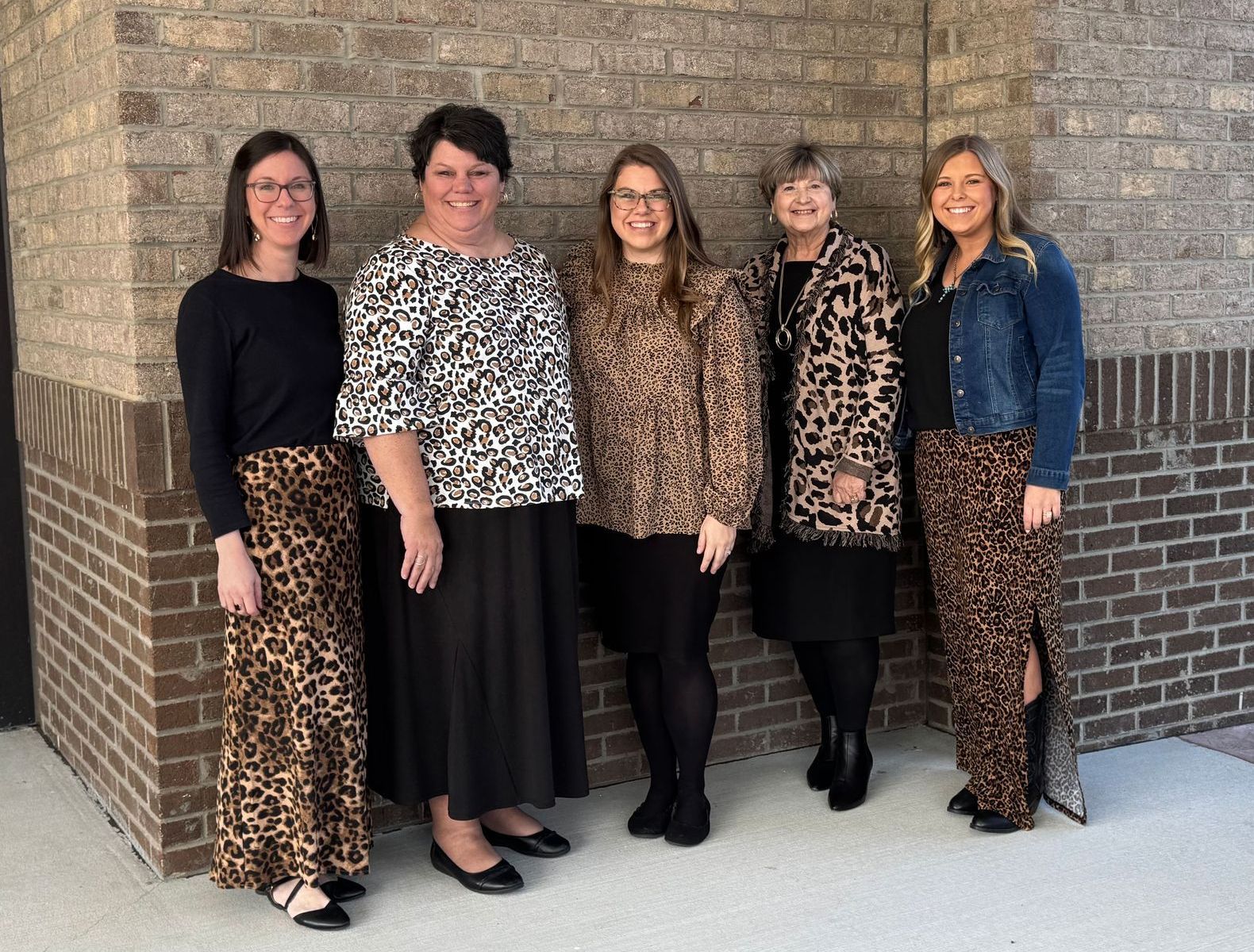 Five women pose by a brick wall; they wear animal print skirts or tops and black clothing.