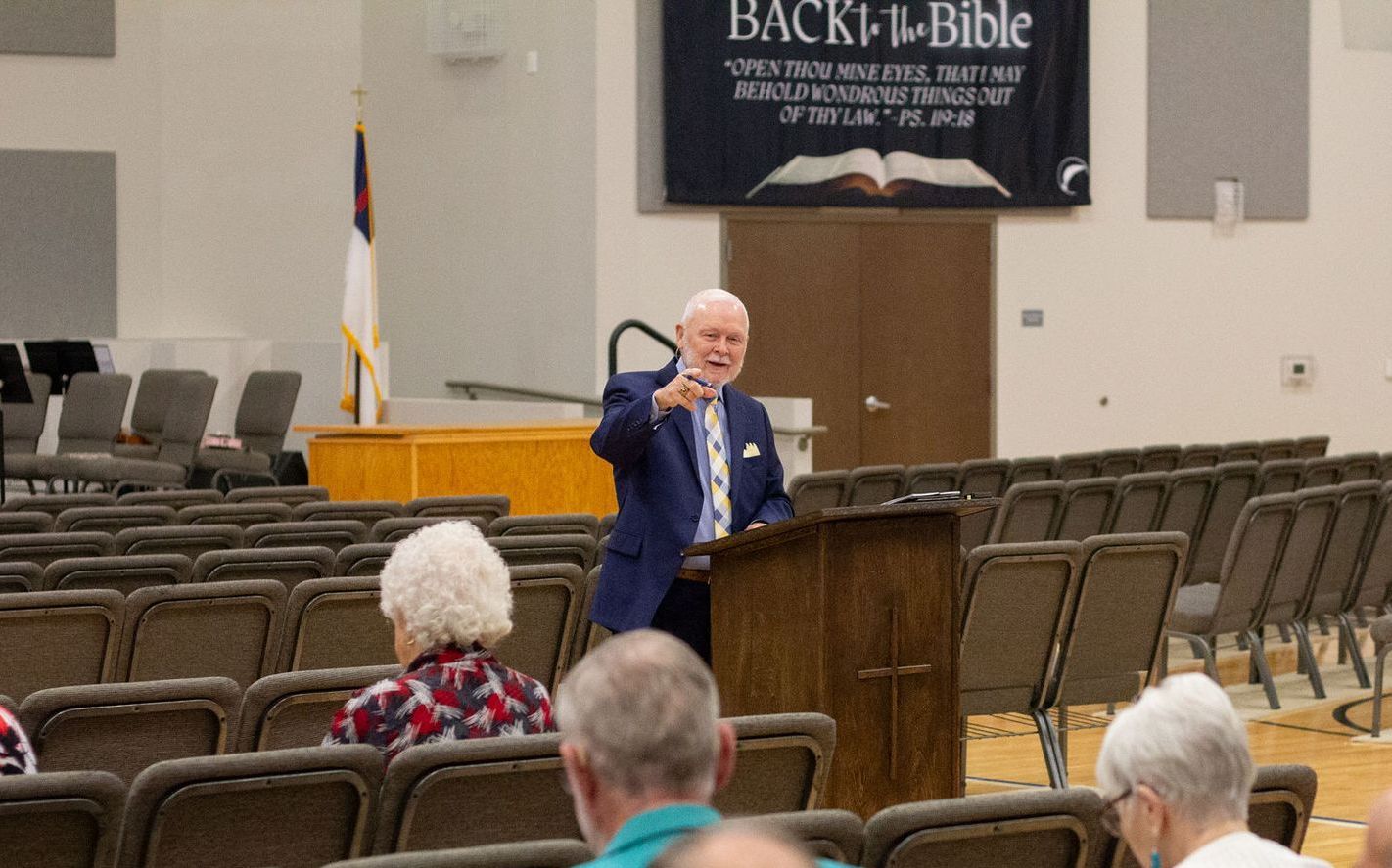 Man in blue suit lecturing from a podium in a church hall, gesturing with his hand.
