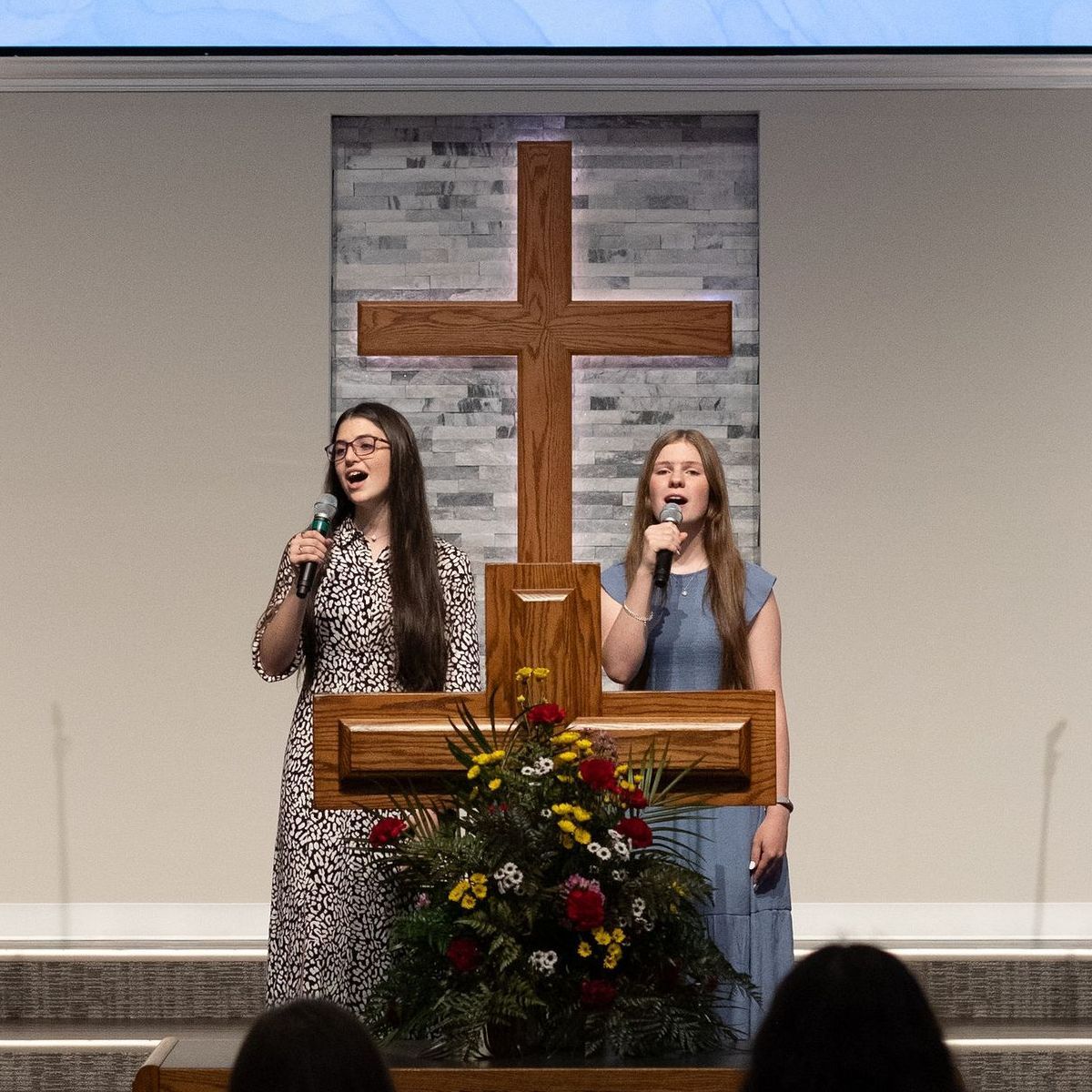 Two women singing into microphones in front of a wooden cross and floral arrangement on a stage.