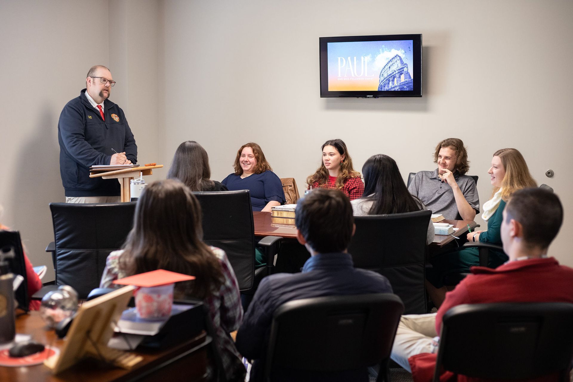 Group meeting: Man speaks to seated attendees. Office setting; TV on wall.