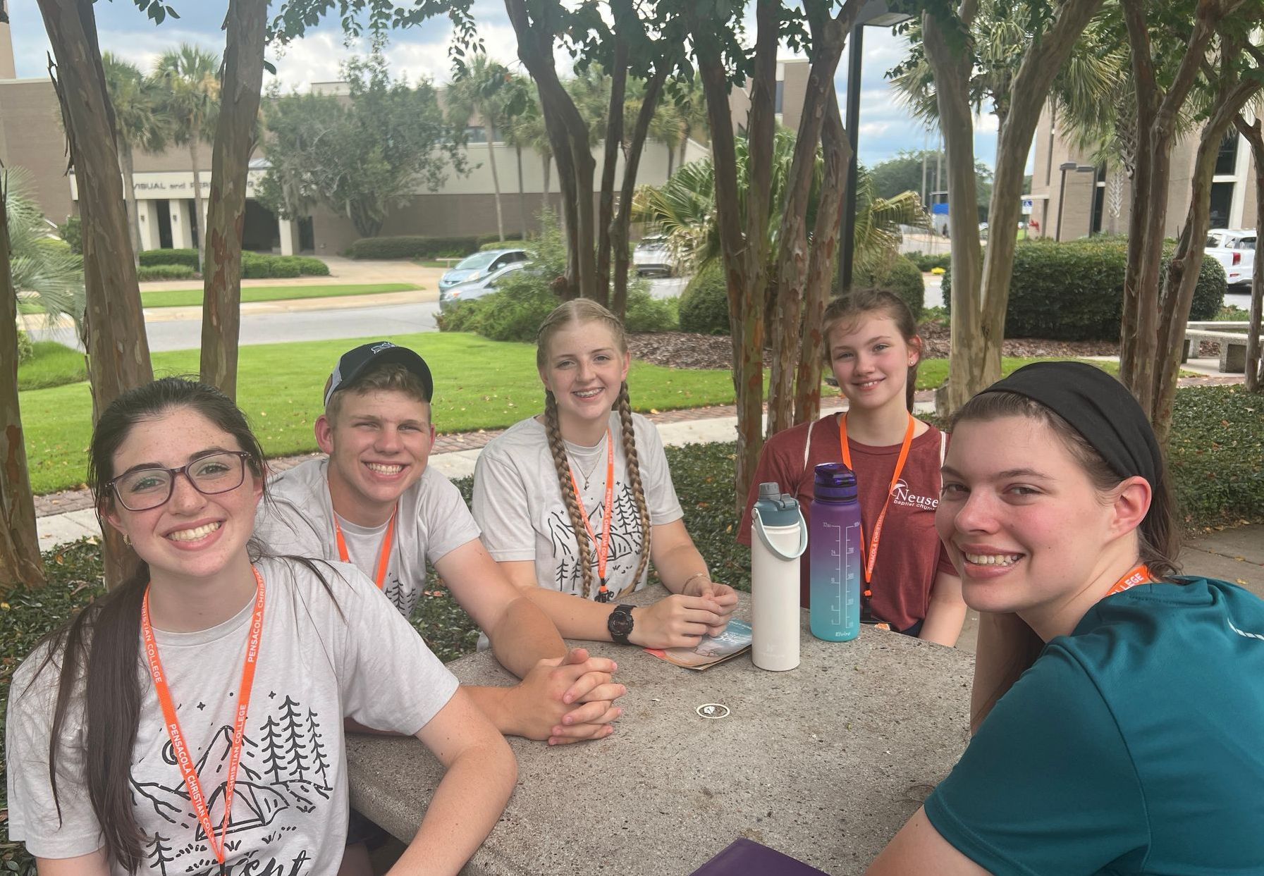 Five young people smile at a table outdoors. They wear lanyards, near trees and a building.