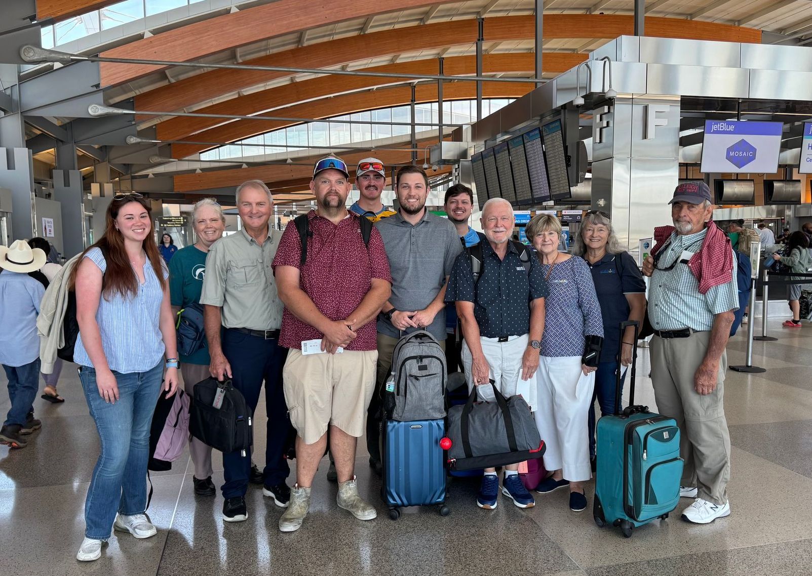 Group of people smiling at an airport, some with luggage, near flight information displays.