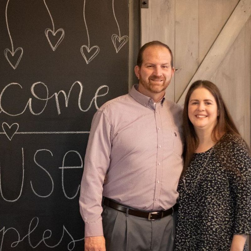 Couple standing in front of a chalkboard sign, man in red shirt, woman in black floral top.