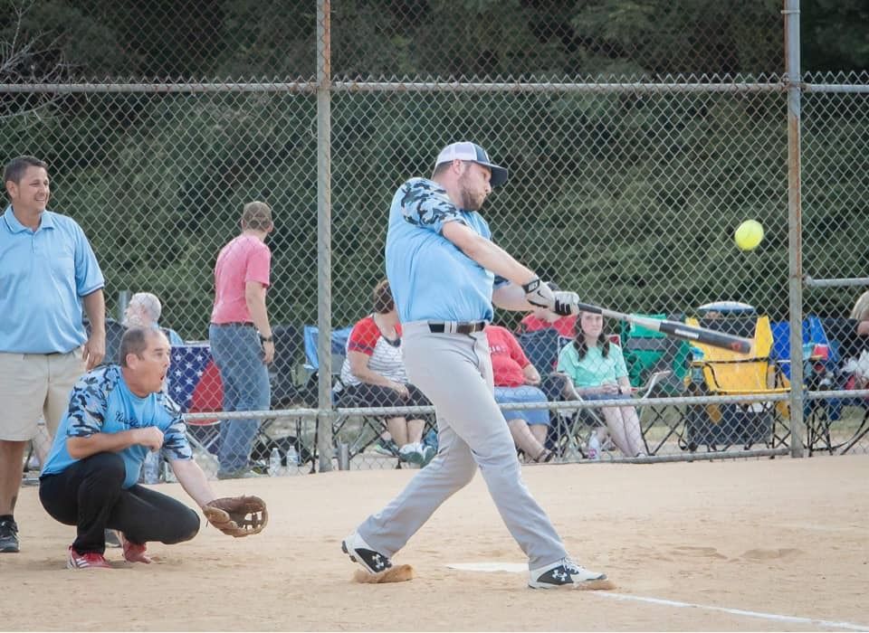 Man in blue shirt swings bat at softball game, umpire watches. Spectators in background.
