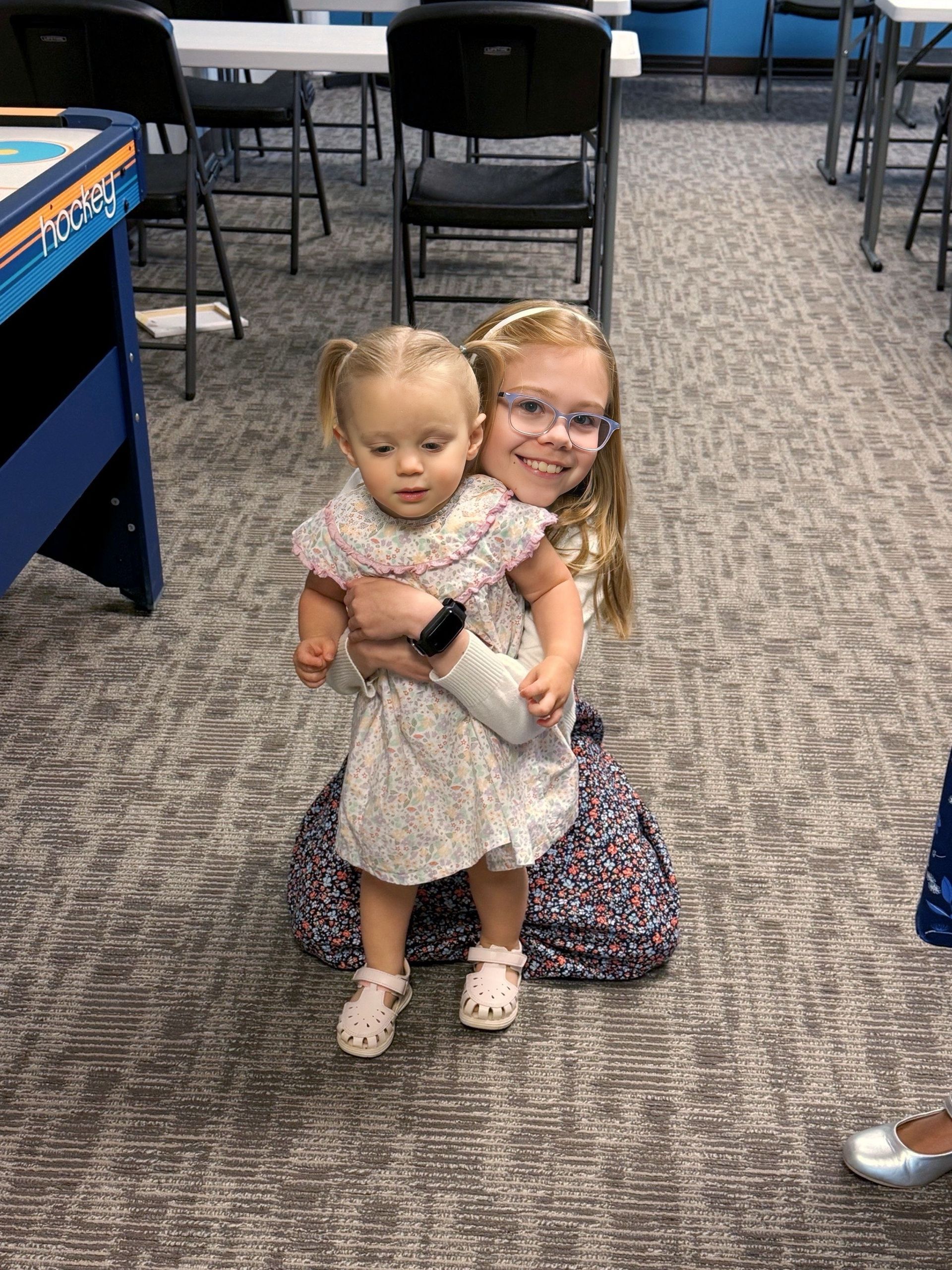 Girl holds a toddler, both smiling in a room with chairs and a table.