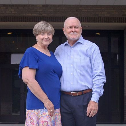 Pastor David and Vickie Shaffer standing in front of a building; woman in blue shirt and skirt, man in blue shirt and slacks.