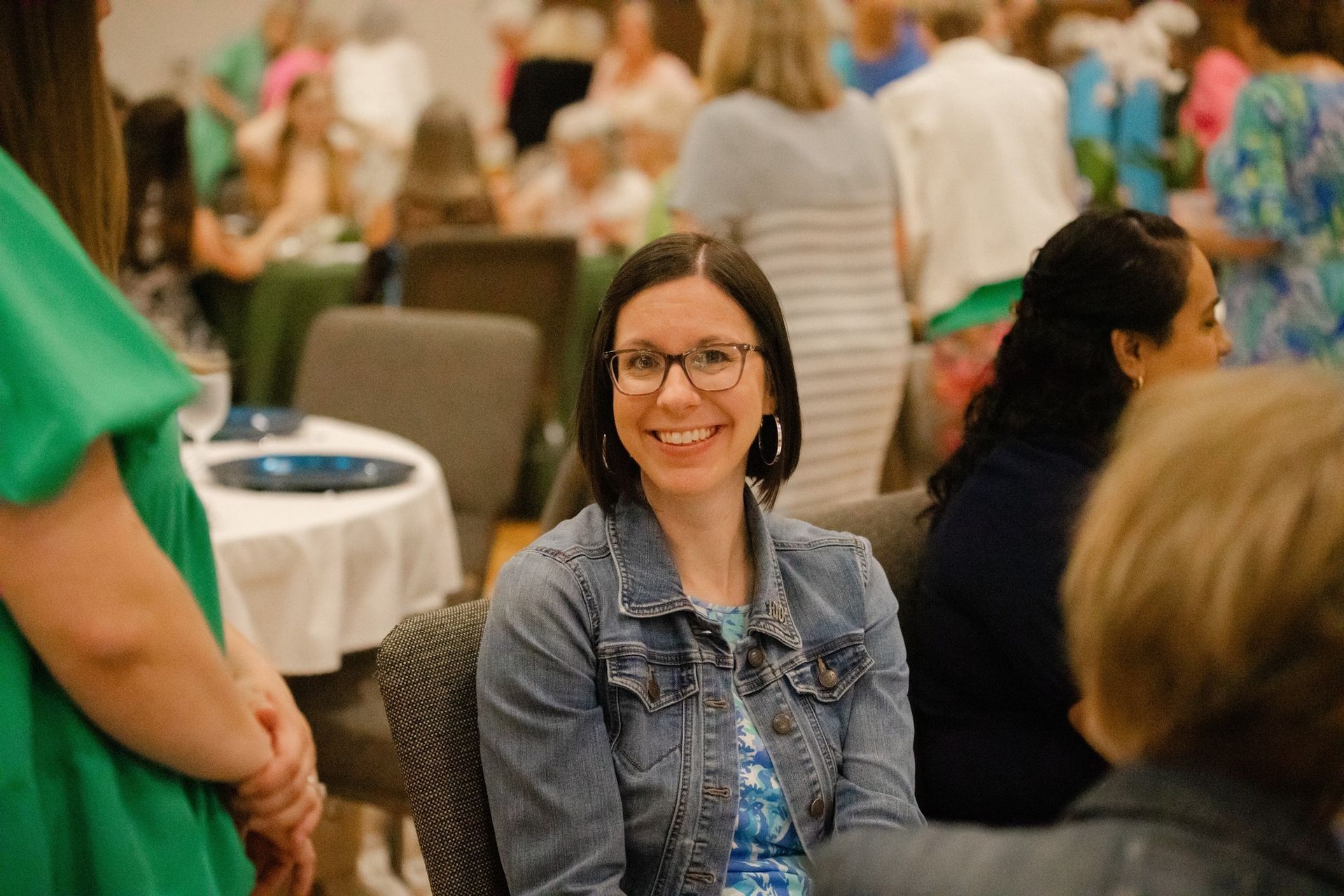 Woman with glasses smiles at an event, wearing a denim jacket and seated at a table.