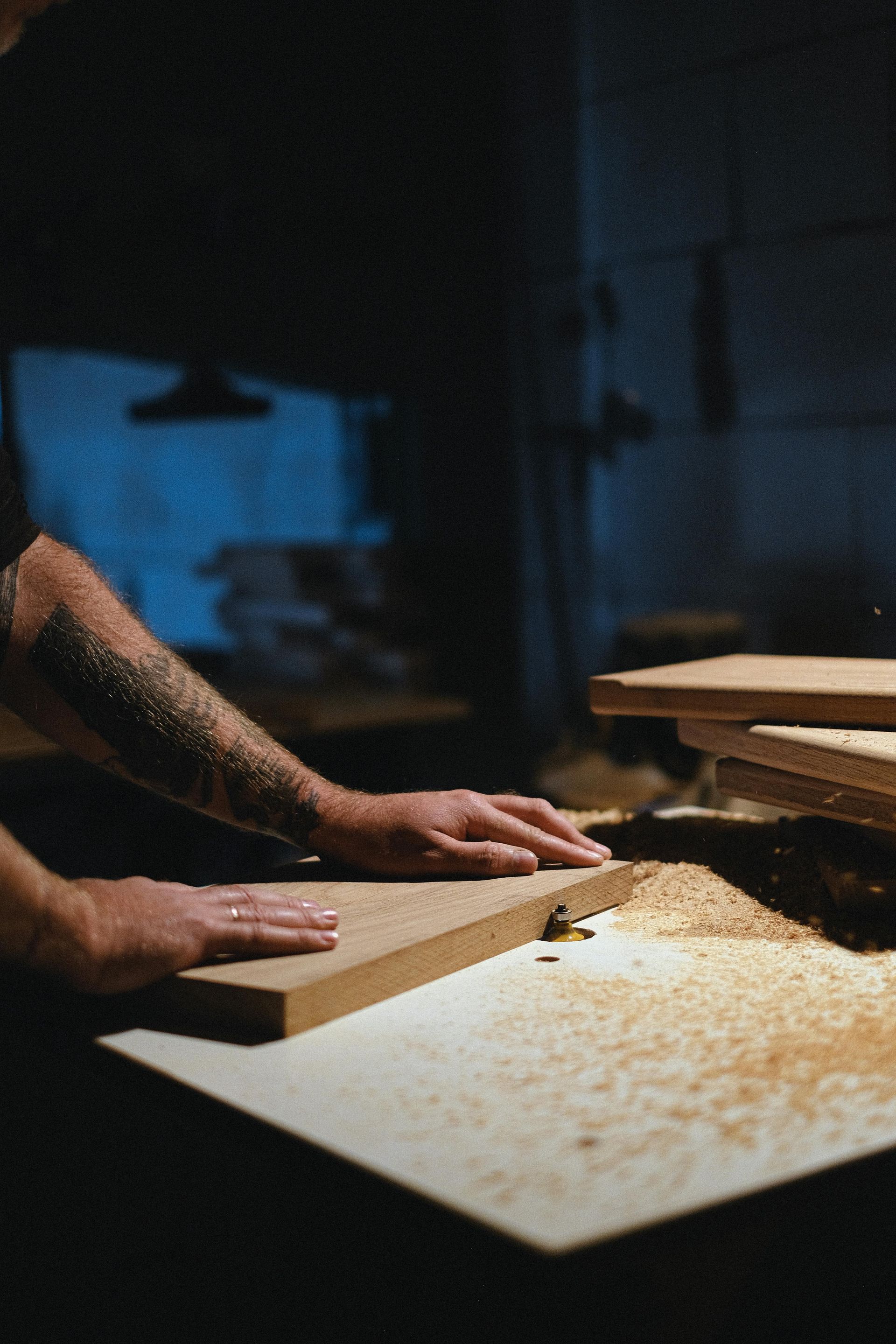 Person using woodworking equipment in a dimly lit workshop, sawdust visible.
