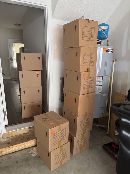 Cardboard moving boxes stacked in a garage doorway, against a wall, and near a water heater.