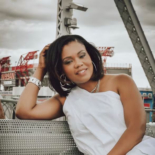 An empty room with cardboard boxes and a clothing rack. Light-colored walls and flooring with natural light coming from a window.