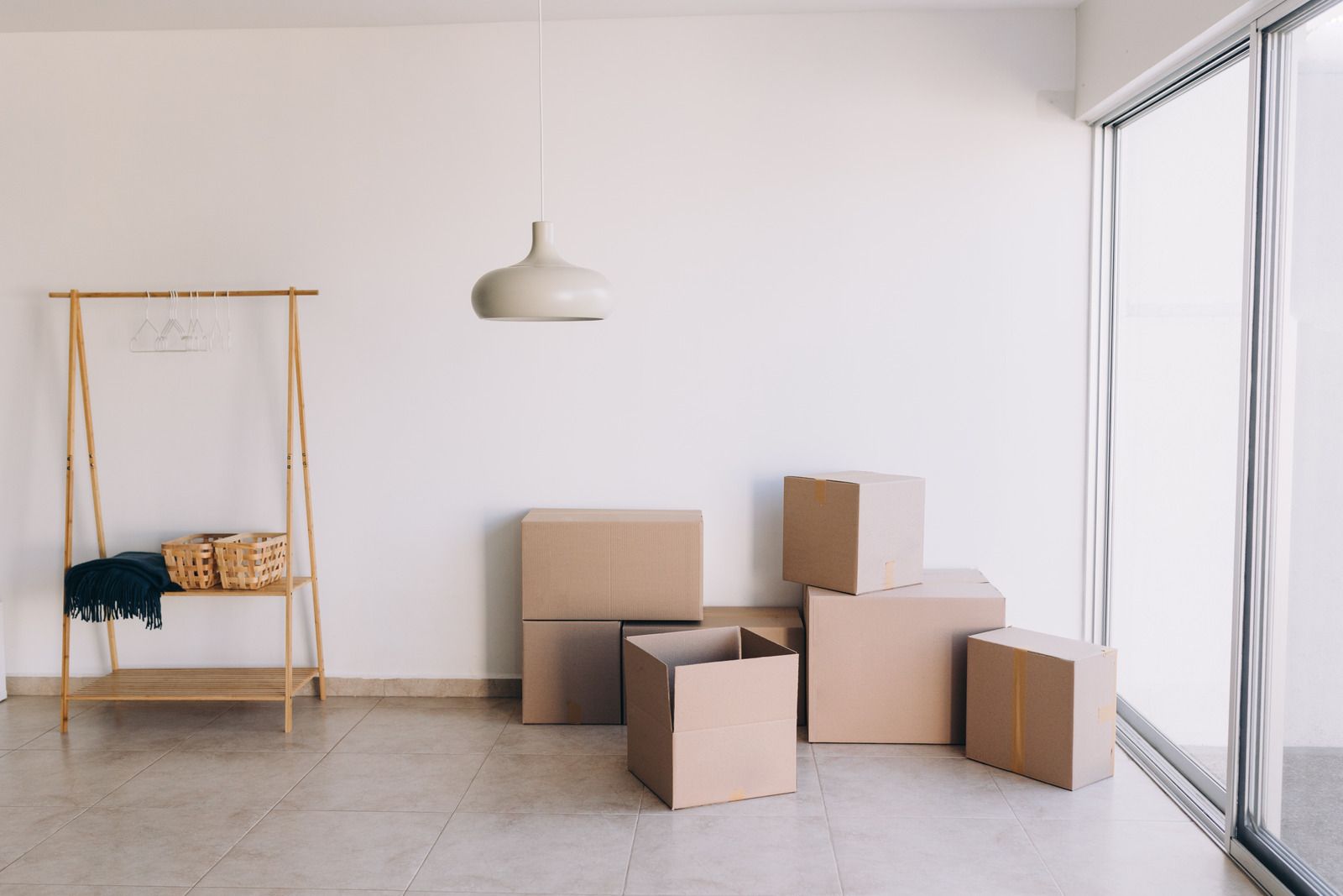 An empty room with cardboard boxes and a clothing rack. Light-colored walls and flooring with natural light coming from a window.