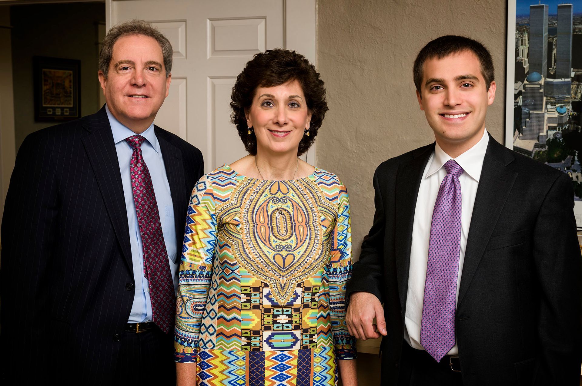 Three formally dressed people smiling indoors. A man on the left and right, woman in the middle.