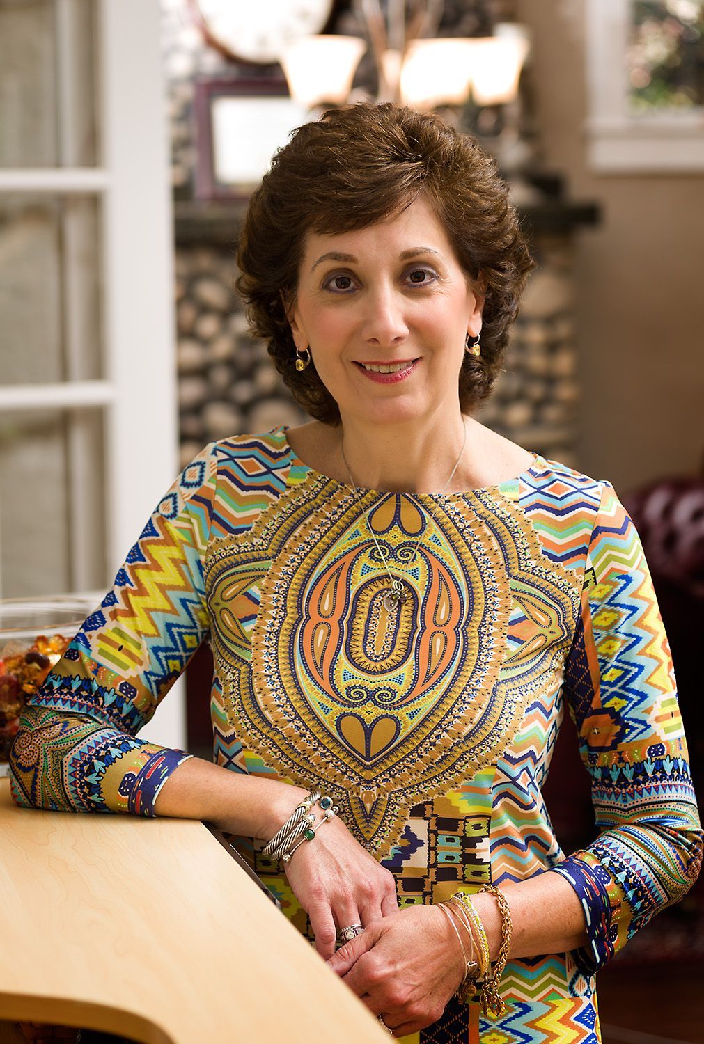 Woman with short brown hair wearing a colorful patterned top, smiling in a well-lit interior.
