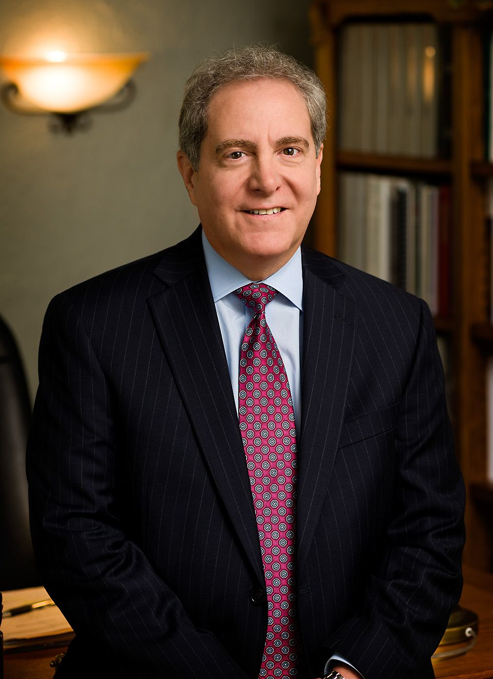 Man in a suit and tie, smiling at the camera. Brown hair, blue shirt, and bookcase visible in the background.