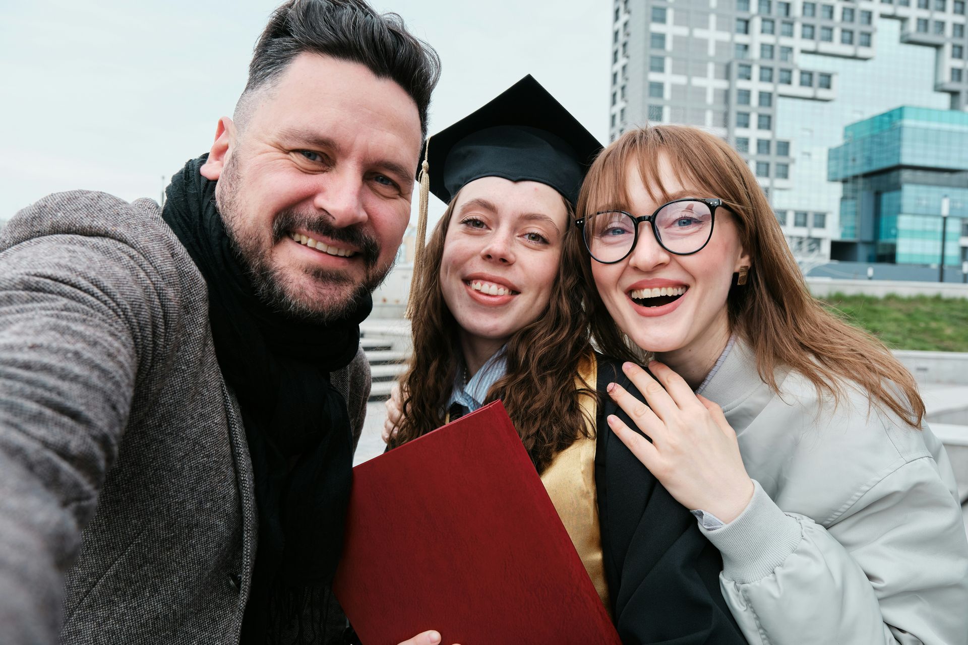 A family takes a selfie; a graduate in cap and gown smiles between her parents, outside a modern building.