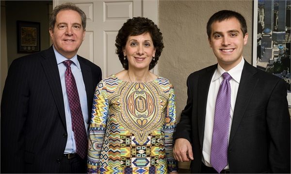 Three formally dressed people smiling indoors. A man on the left and right, woman in the middle.