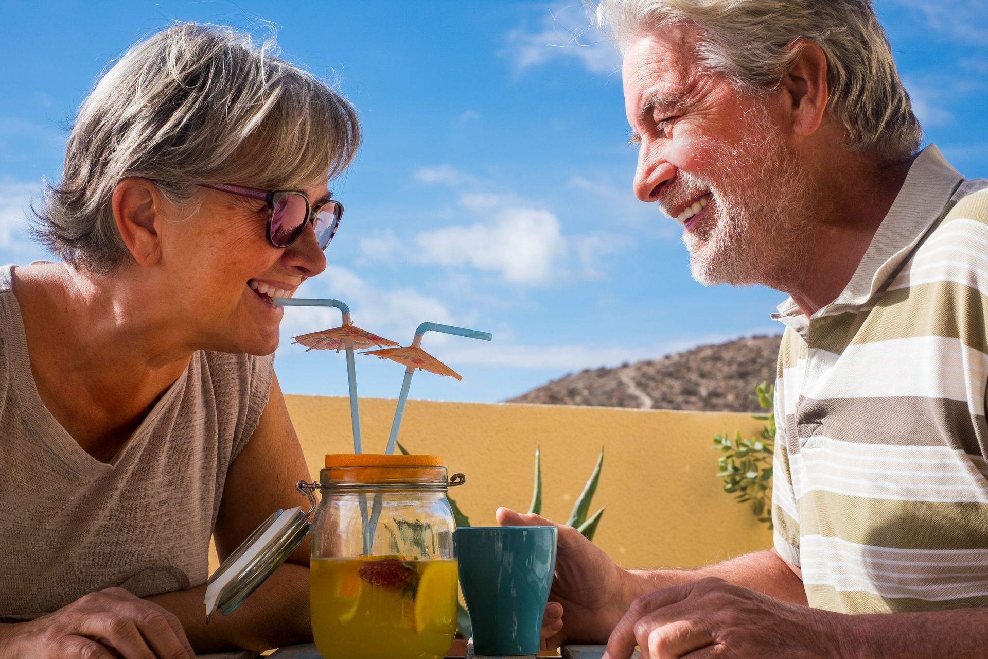 Elderly couple smiles at each other outdoors. They share a drink with straws, set against a sunny sky.
