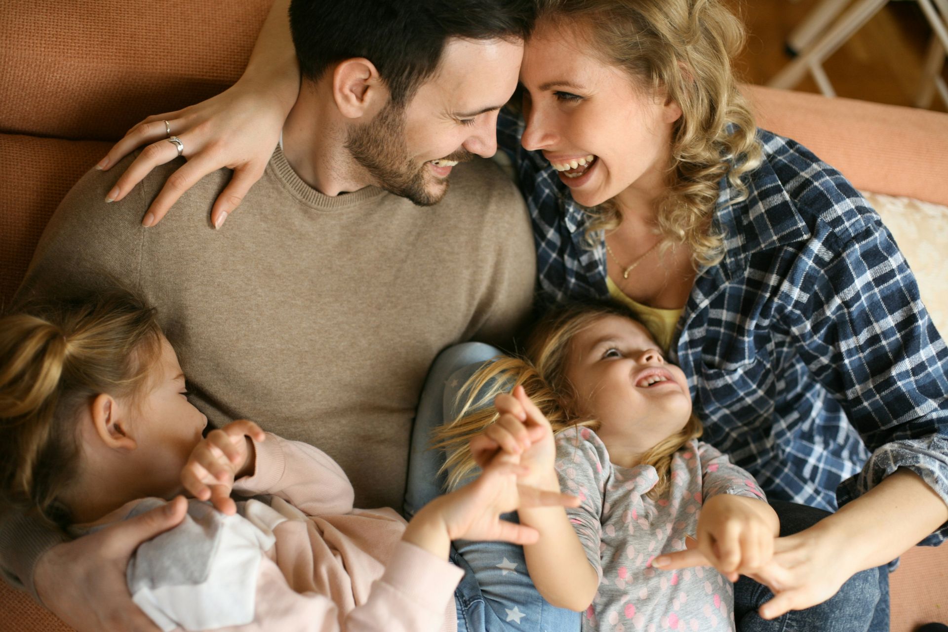 Family of four laughing and embracing on a couch. Two young girls play and giggle with their parents.