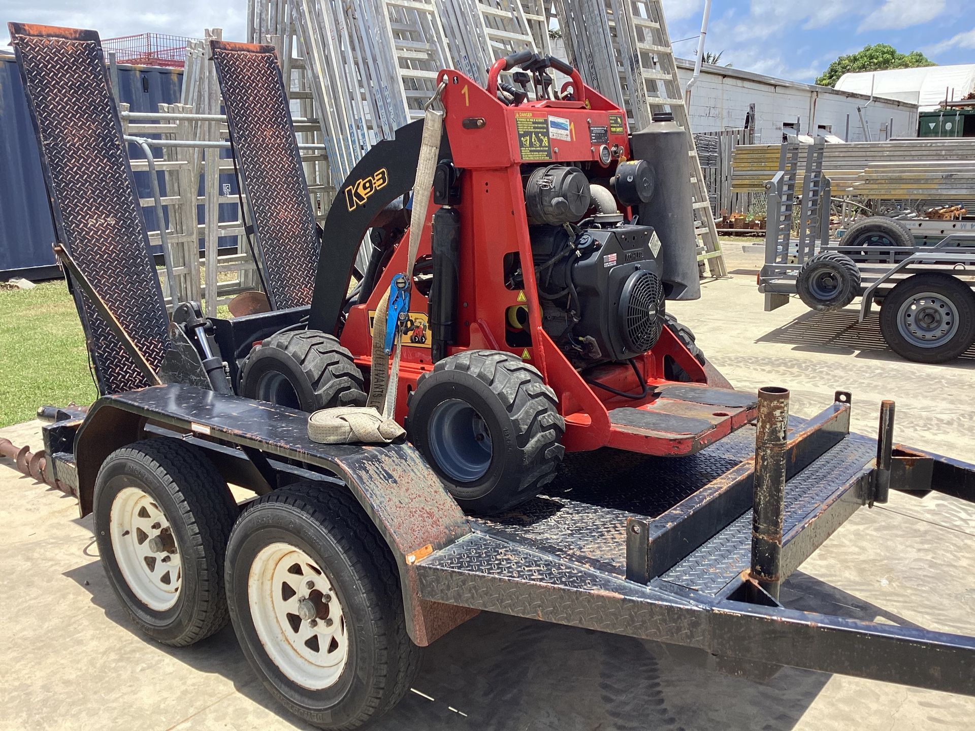 Red Skid Steer Loader Strapped on a Black Flatbed Trailer in an Equipment Yard — Bowen Hire in Bowen, QLD