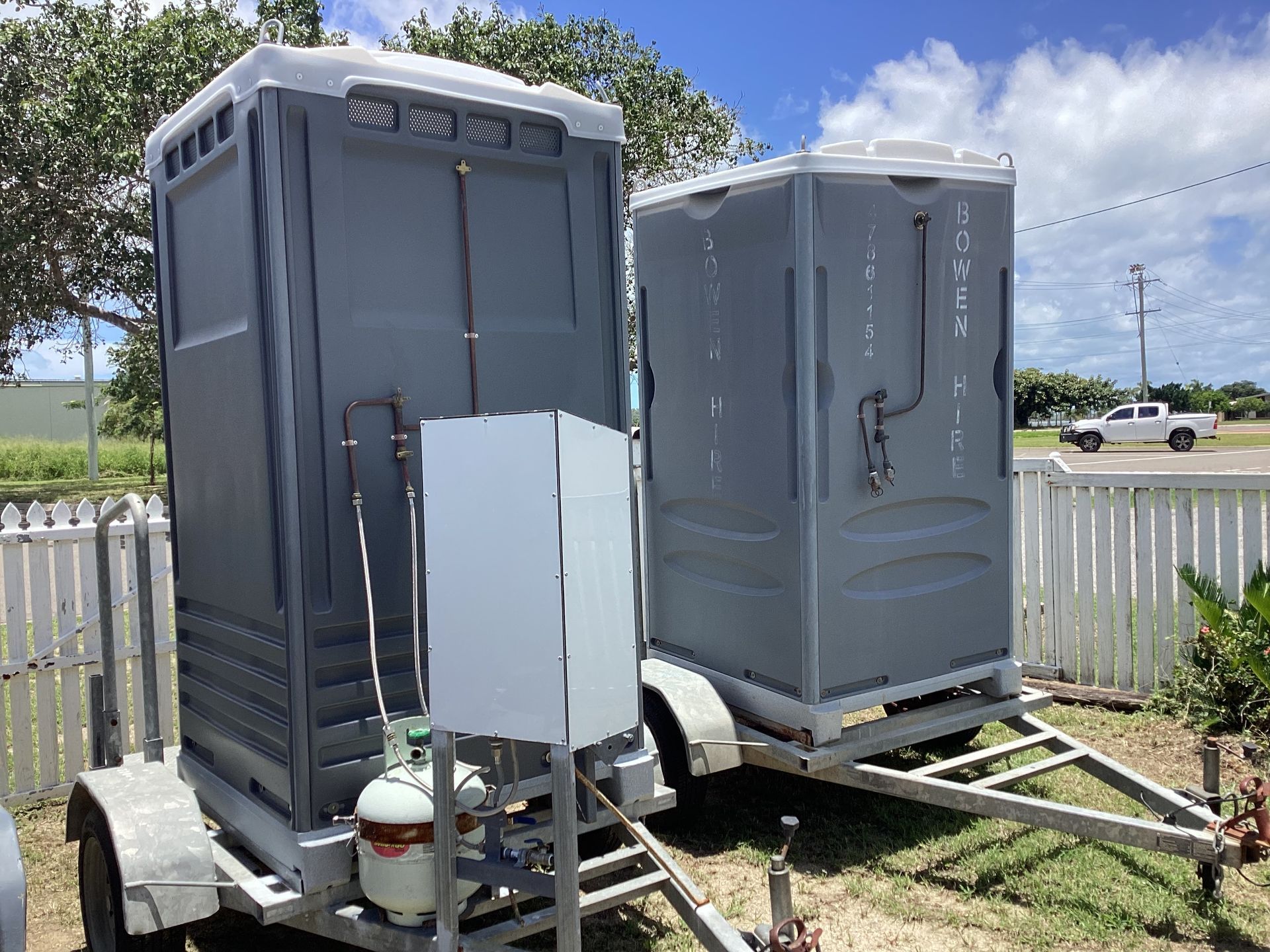 Two Gray Portable Toilets on Trailers in A Grassy Lot by A White Fence— Bowen Hire in Airlie Beach, QLD