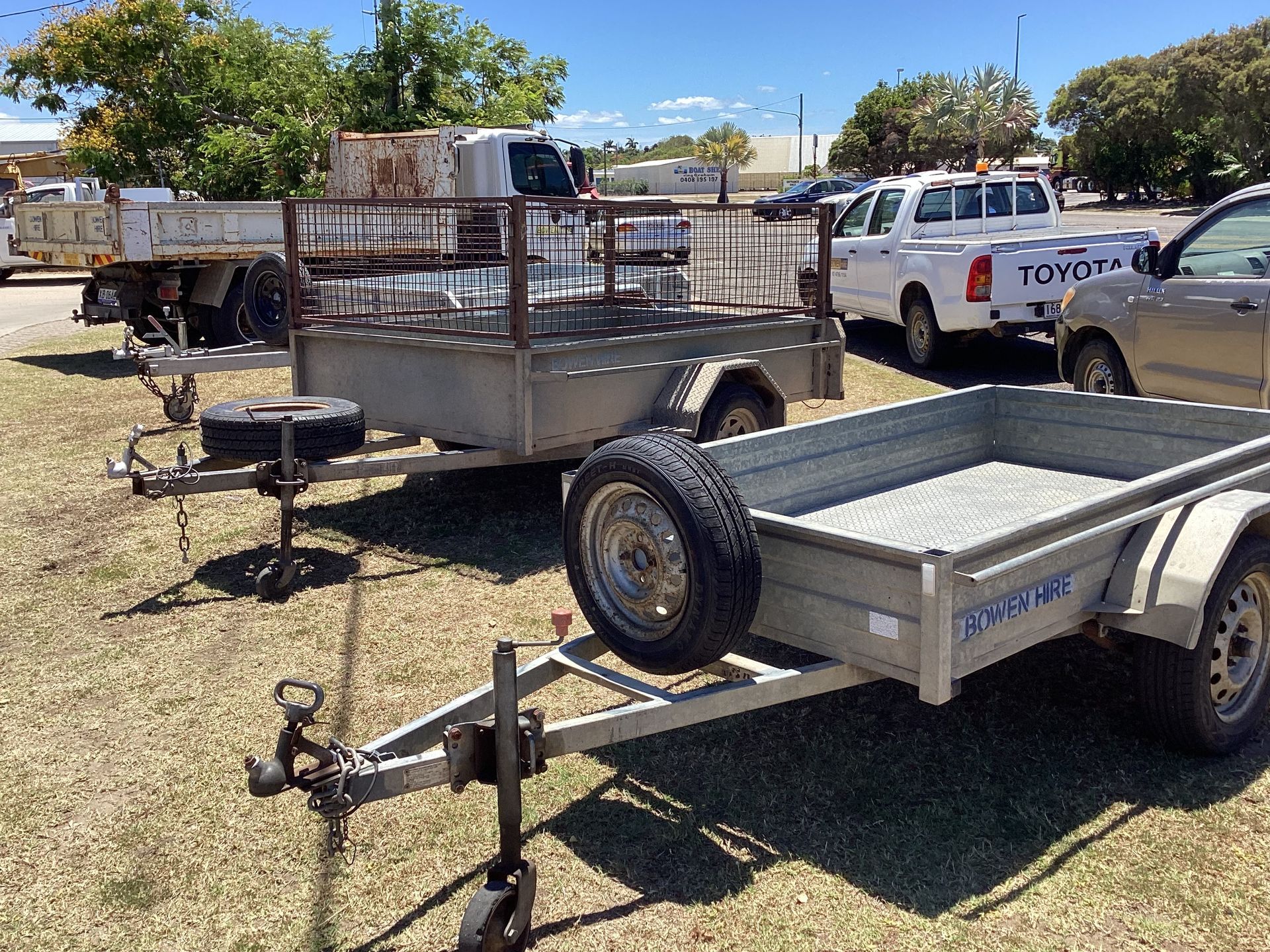 Two Utility Trailers Parked in a Sunny Gravel Lot Near a White Pickup Truck and Trees — Bowen Hire in Bowen, QLD