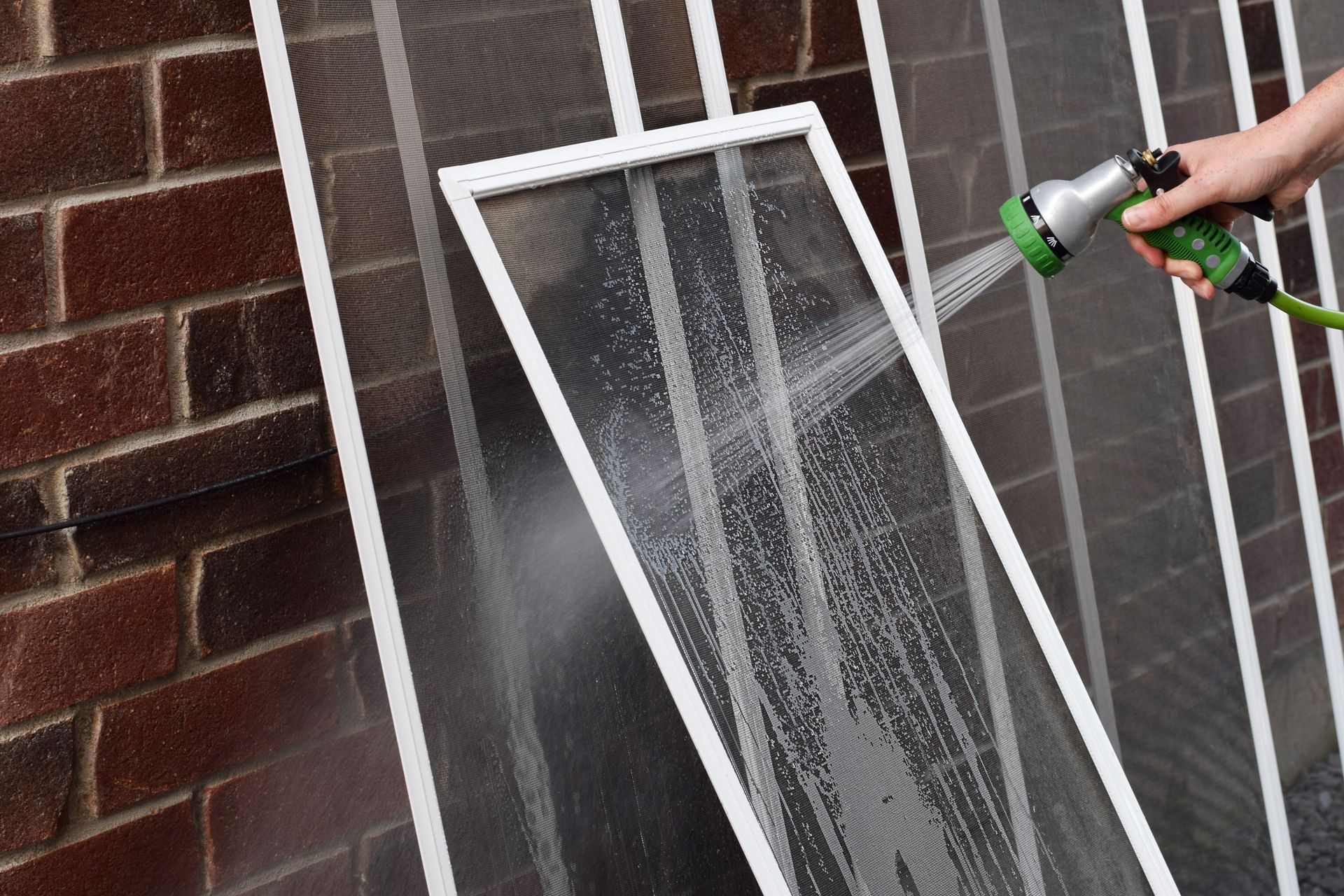 Person spraying water onto white-framed window screens leaning against a brick wall.
