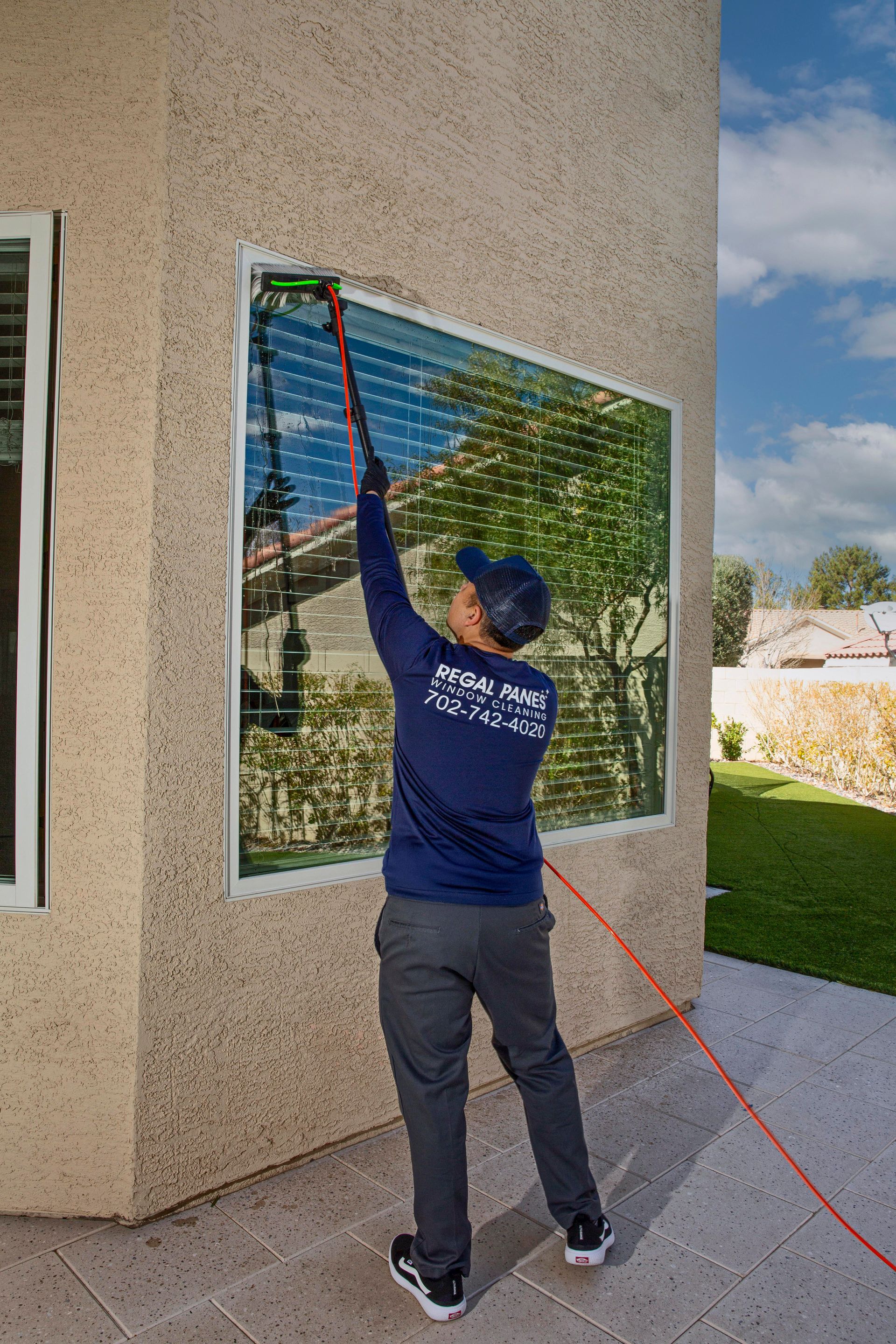 Person washing a window with an extended cleaning tool on a house exterior.